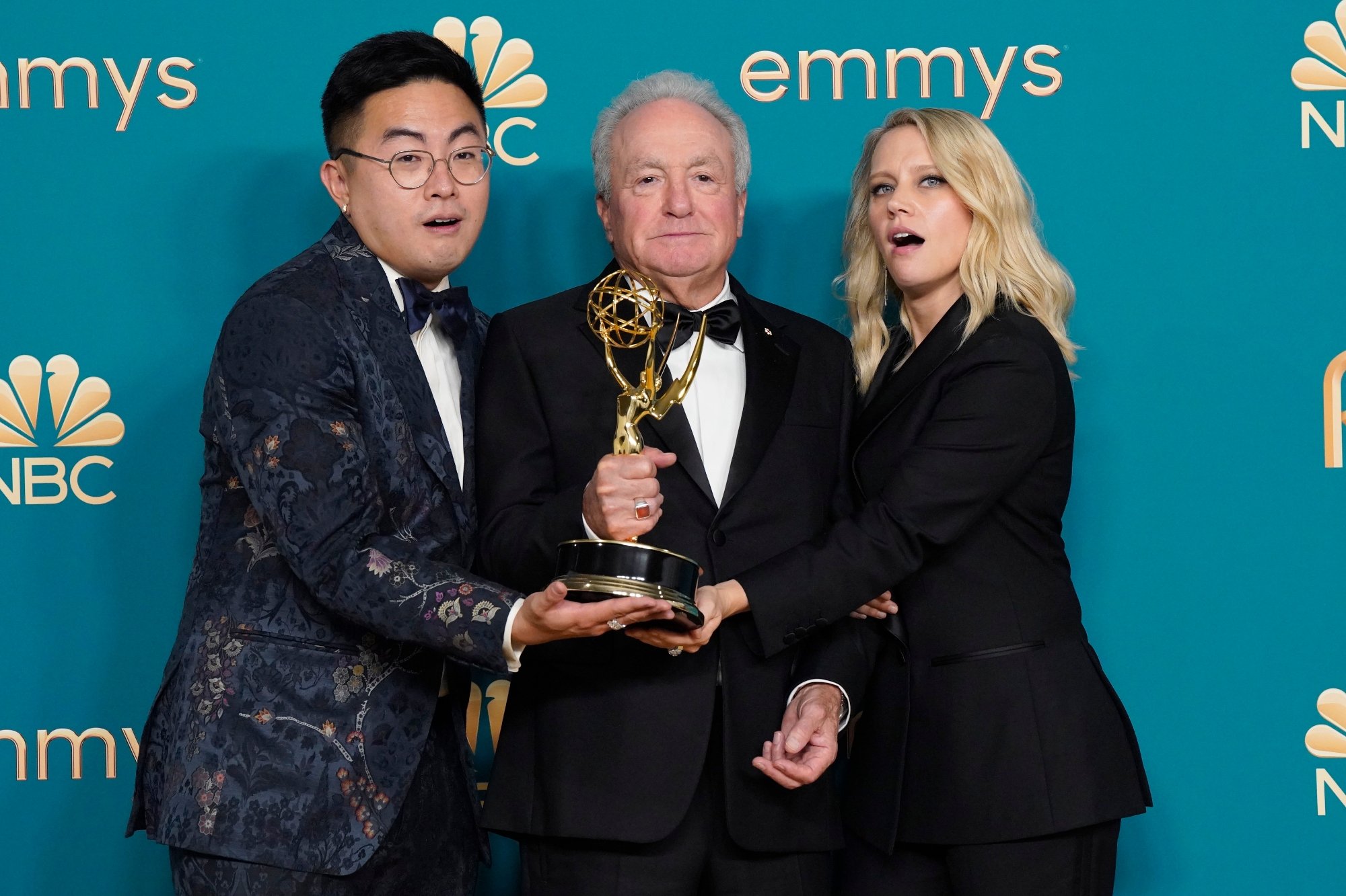 'Saturday Night Live' Bowen Yang, Lorne Michaels, and Kate McKinnon standing in front of Emmys step and repeat. They're all holding an Emmy award with their mouths slightly open in excitement.