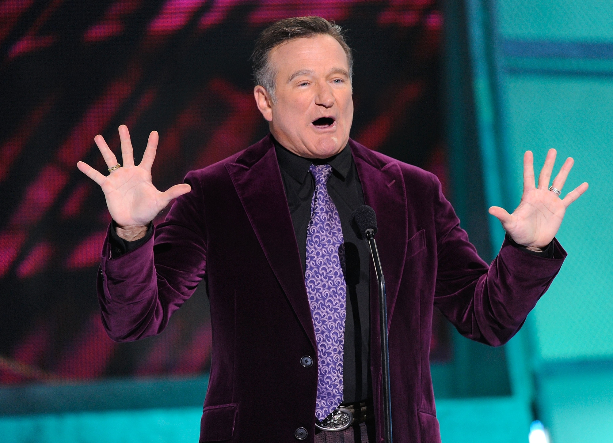 'Saturday Night Live' host Robin Williams. He's wearing a purple velvet suit jacket and a purple tie. He's holding his hands up with a look of surprise on his face.
