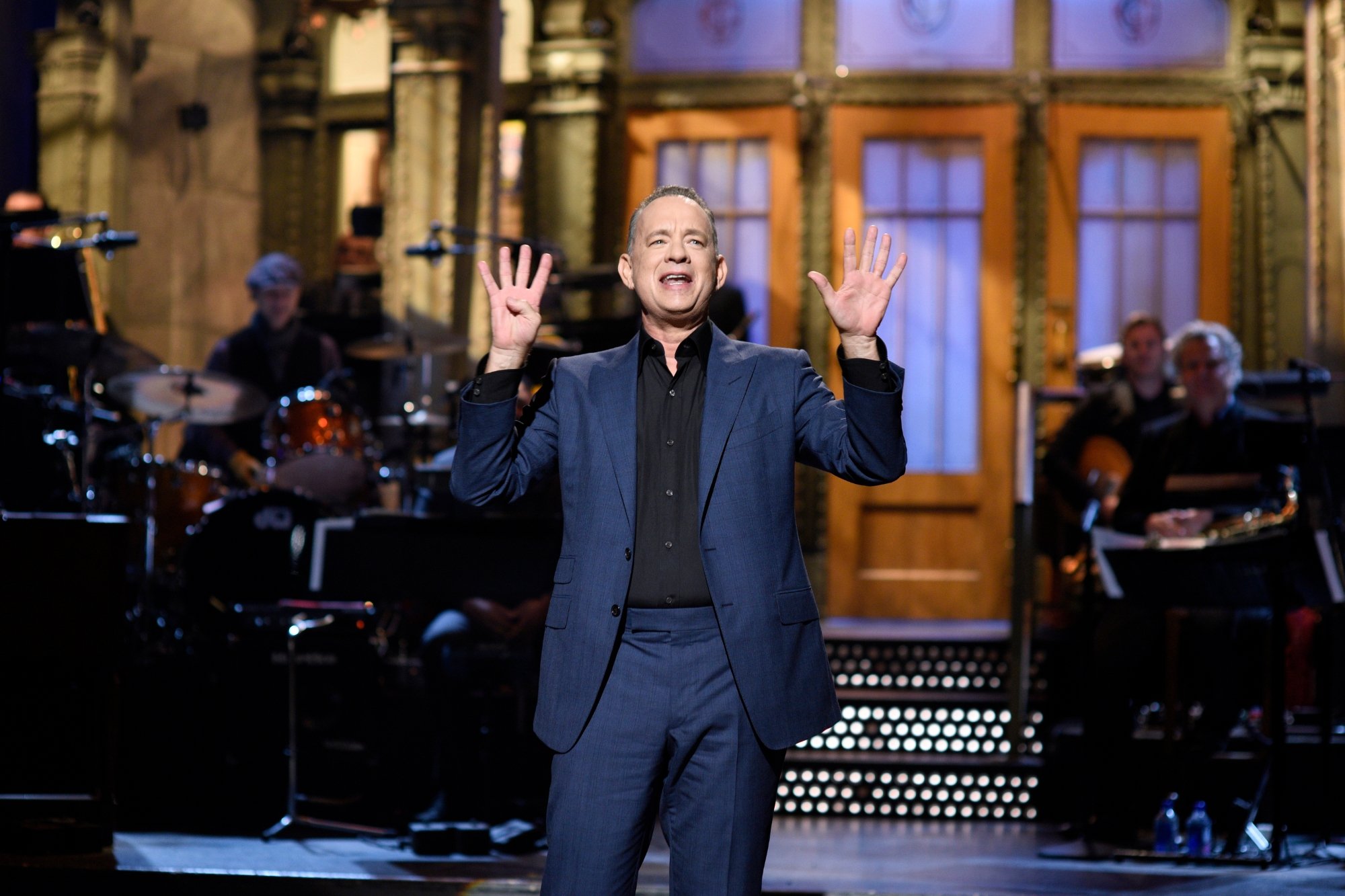 'Saturday Night Live' host Tom Hanks holding up 9 fingers, wearing a navy blue suit and a black collared shirt on the stage.