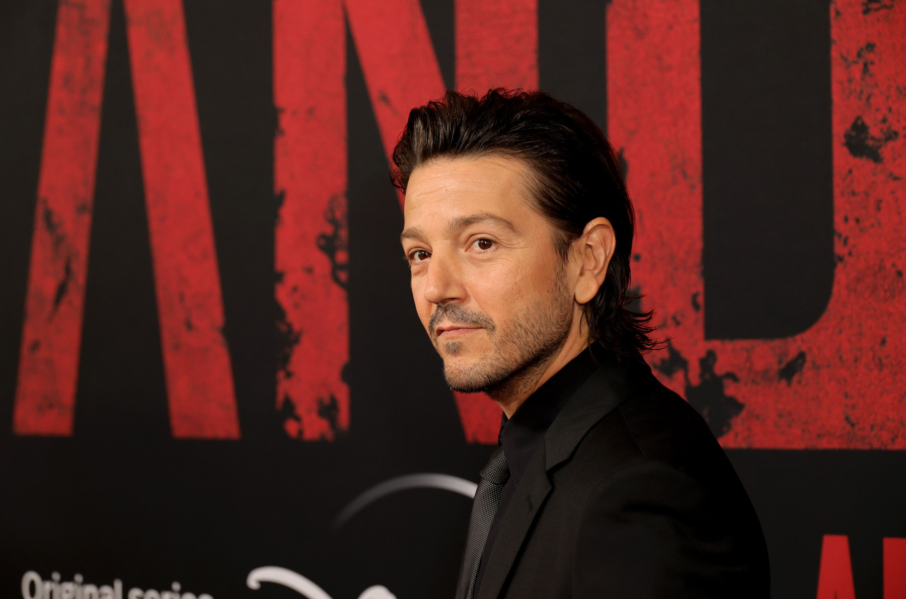 Diego Luna, who headlines 'Star Wars' series 'Andor' on Disney+. In the image, he's standing in front of a wall with the show's logo, wearing a black suit, and has his hair slicked back.