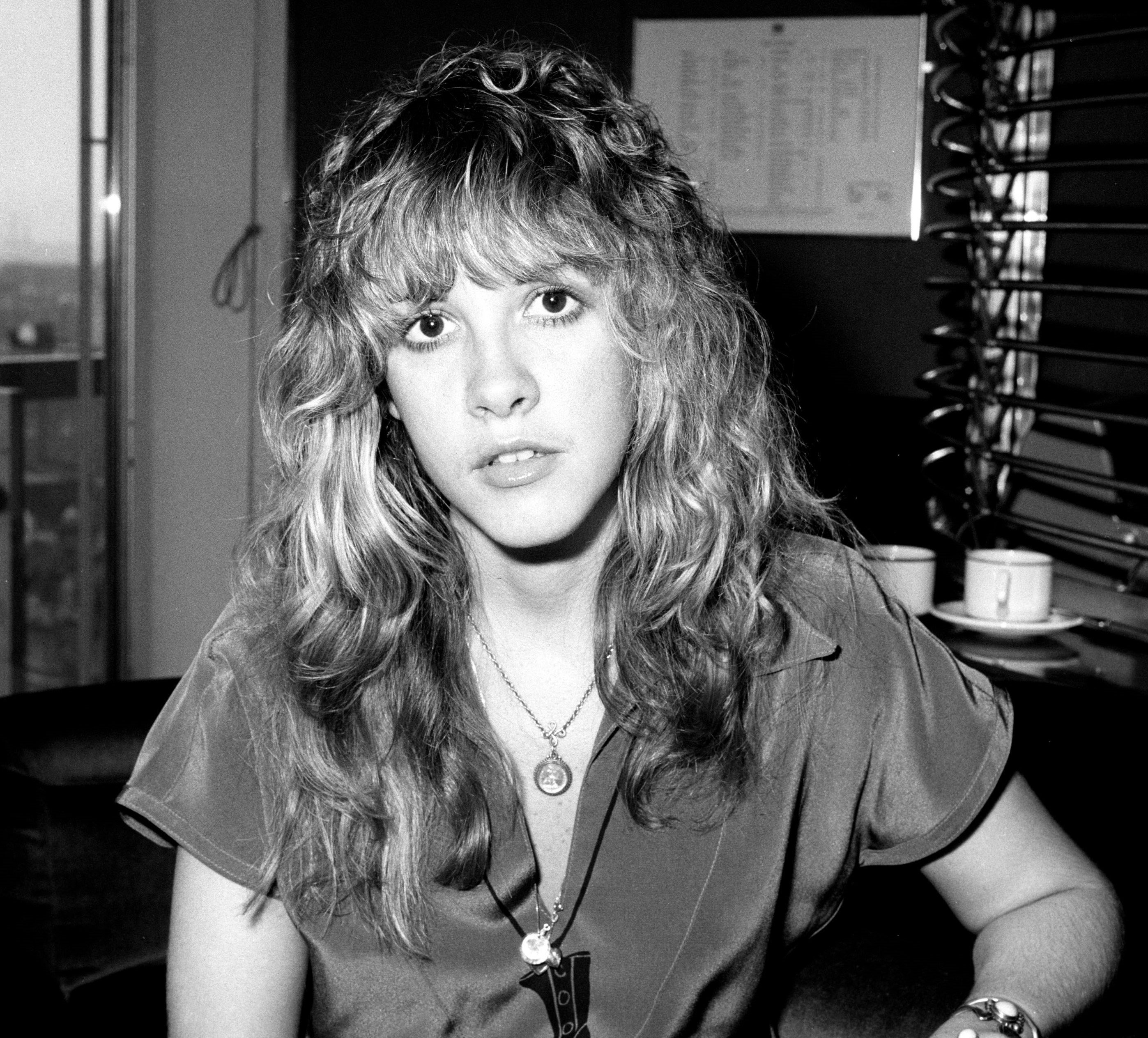A black and white picture of Stevie Nicks sitting near a window and a table. 