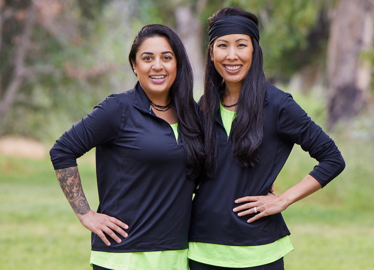 Aastha Lal and Nina Duong on 'The Amazing Race' Season 34 stand next to each other in match green shirts and black jackets.