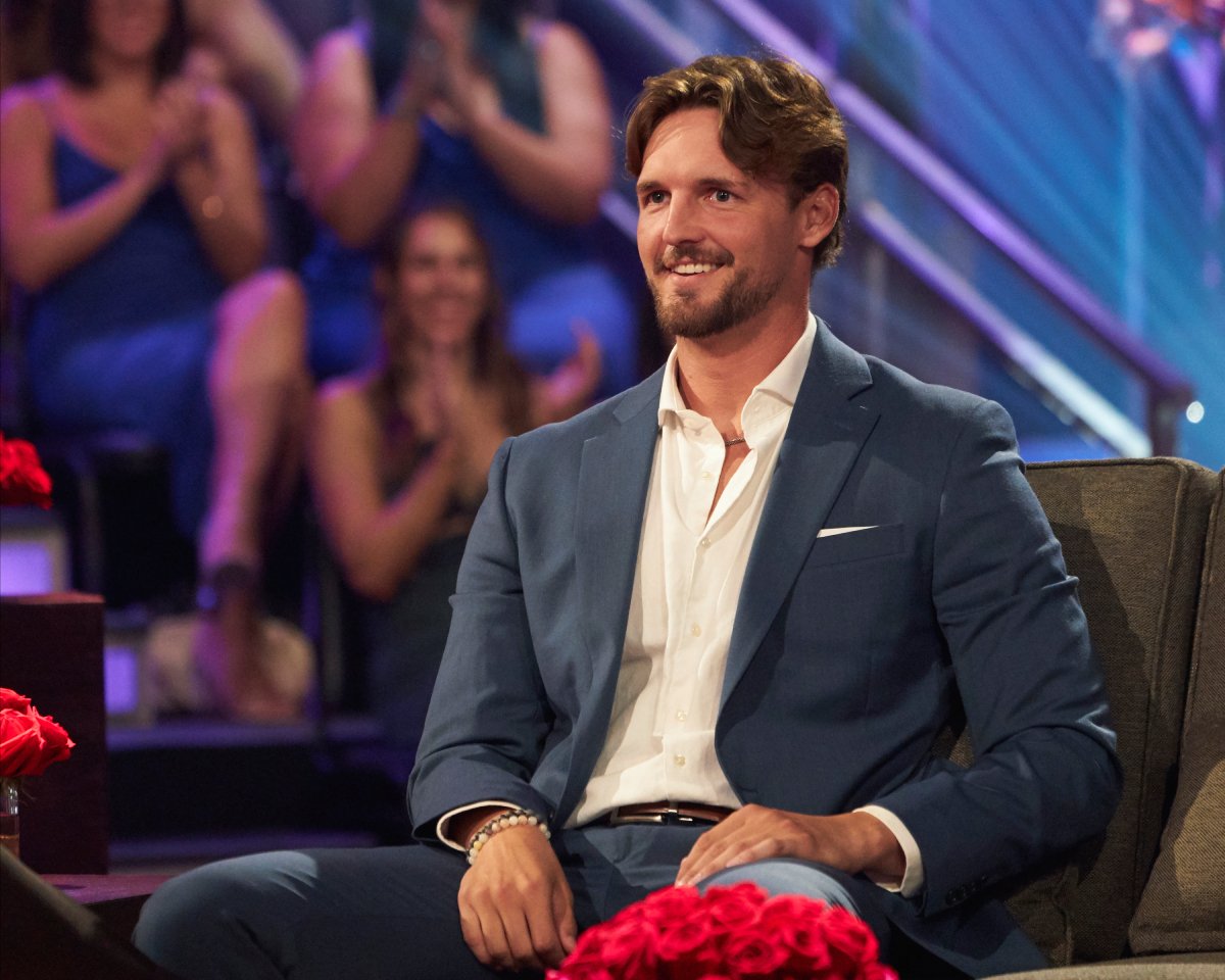 Logan Palmer attends The Bachelorette 2022 Men Tell All wearing a blue/grey suit jacket and white button-down shirt. 