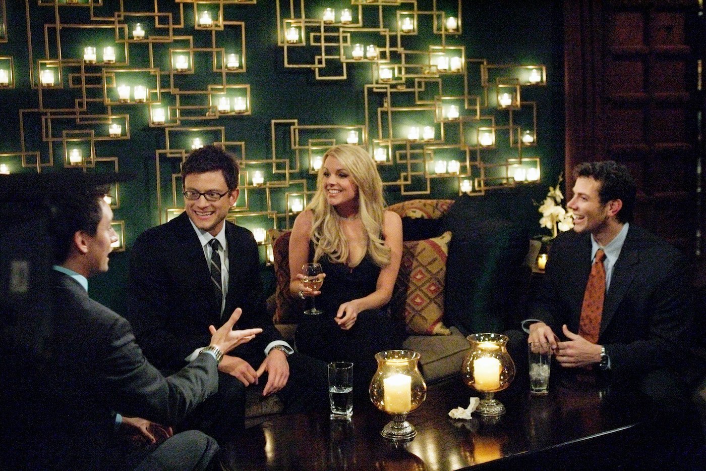Ali Fedotowsky sits on a couch surrounded by three men wearing suits