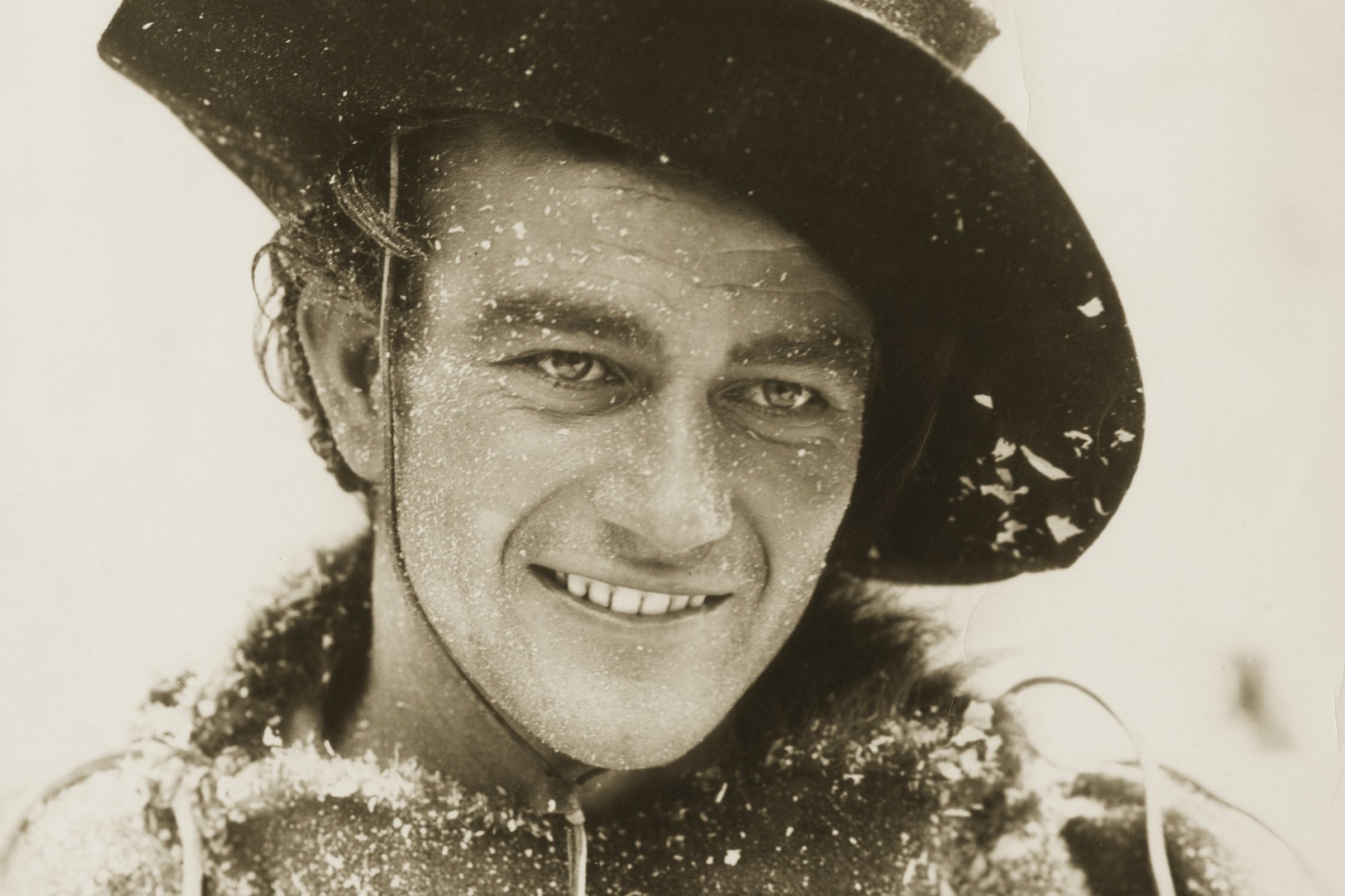 'The Big Trail' movie star John Wayne. He's wearing a cowboy hat and Western costume. Wayne has a smile on his face in a sepia tone picture.
