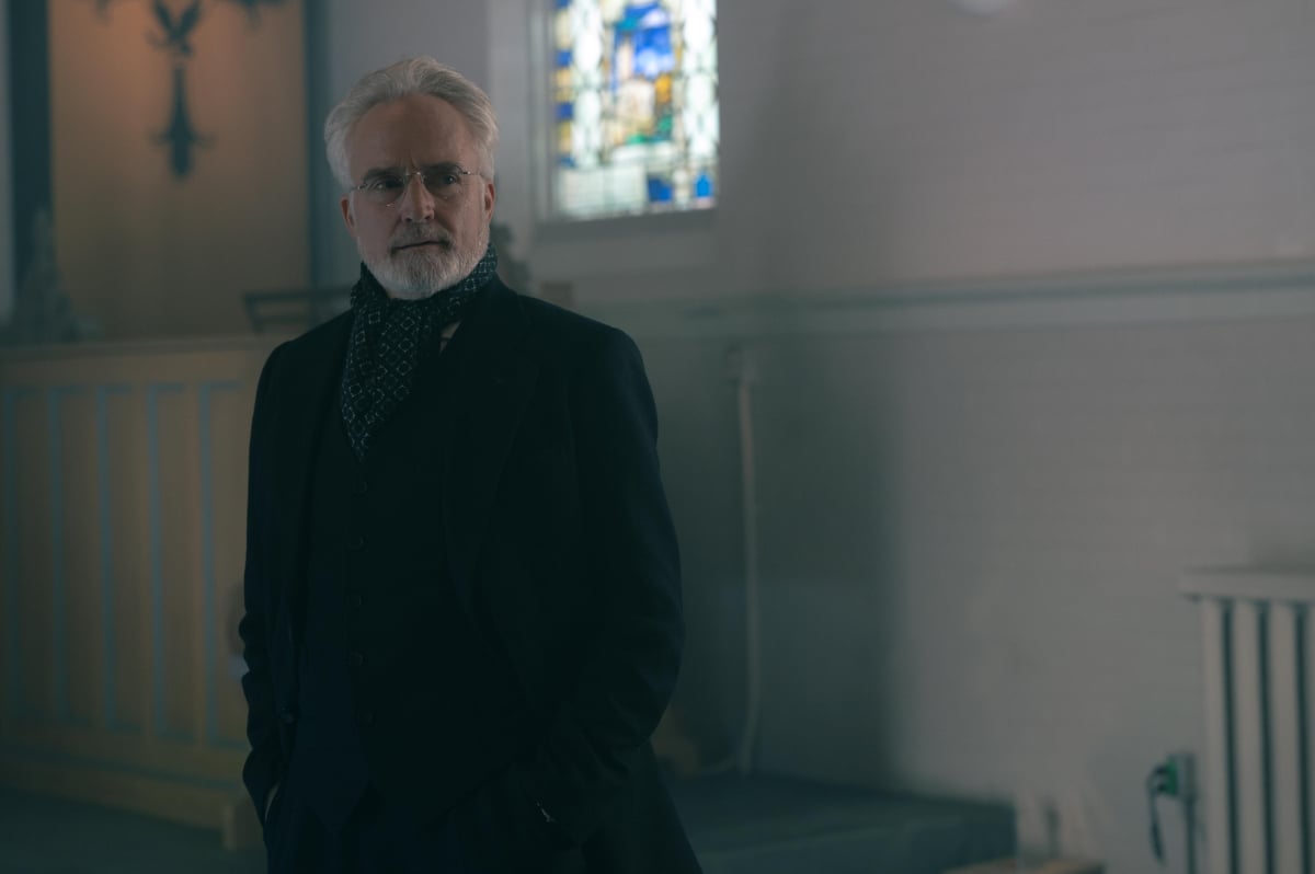 Bradley Whitford as Commander Lawrence in The Handmaid's Tale. Commander Lawrence stands in a church wearing a black coat and a scarf.