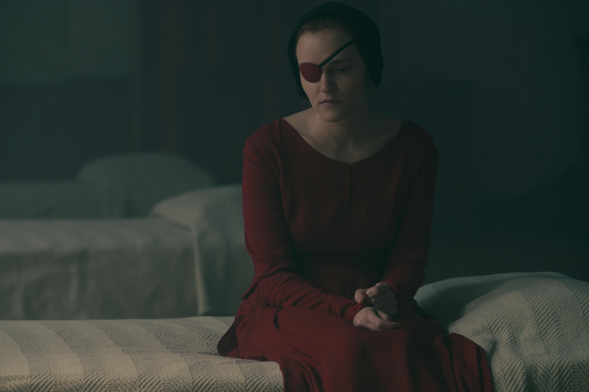 Madeline Brewer as Janine in The Handmaid's Tale Season 5. Janine sits on a bed at the Red Center wearing a handmaid's red dress and an eye patch.