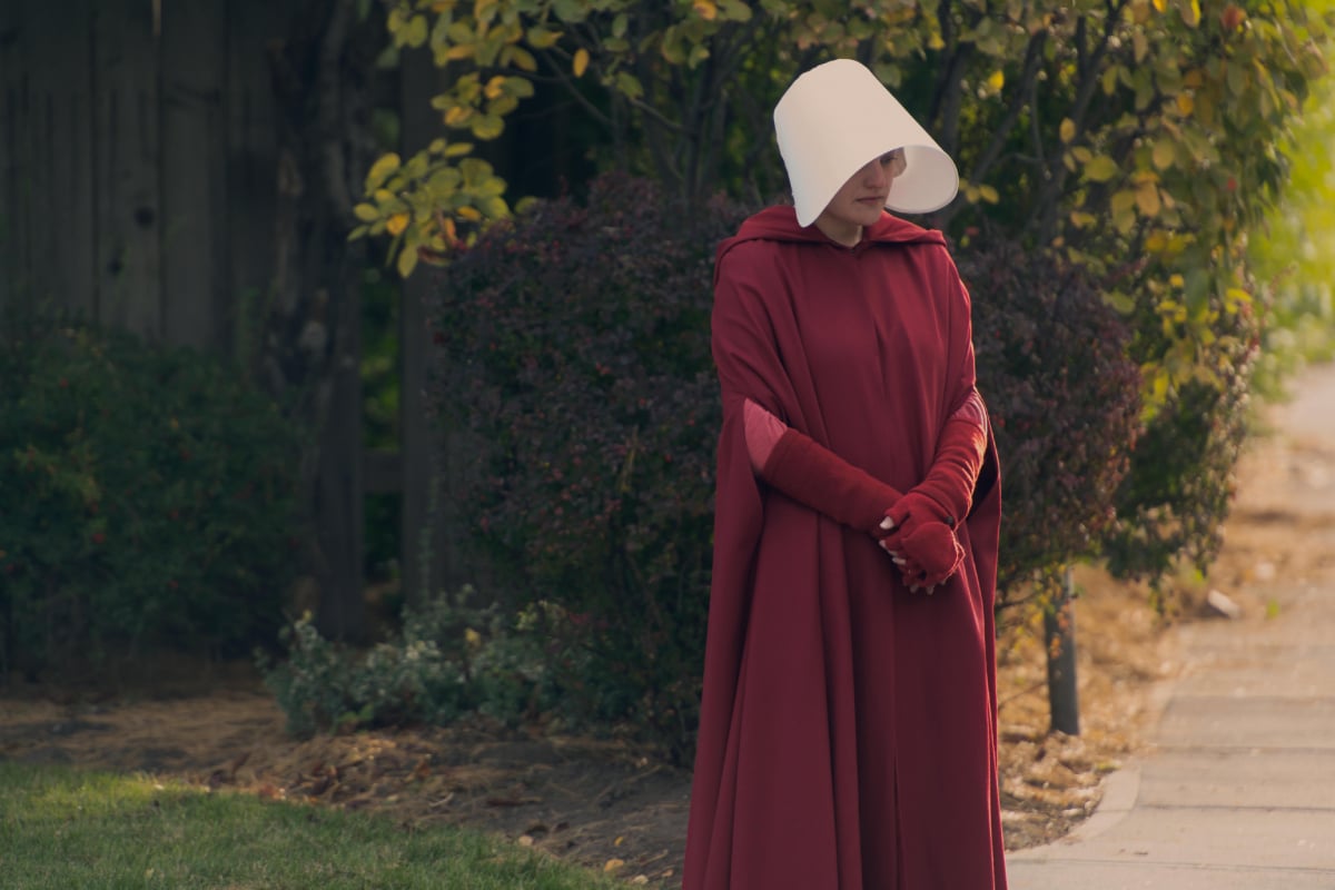 The Handmaid's Tale has been renewed for season 6. Elisabeth Moss as June stands outside in red cloak with the white blinder on her head. 