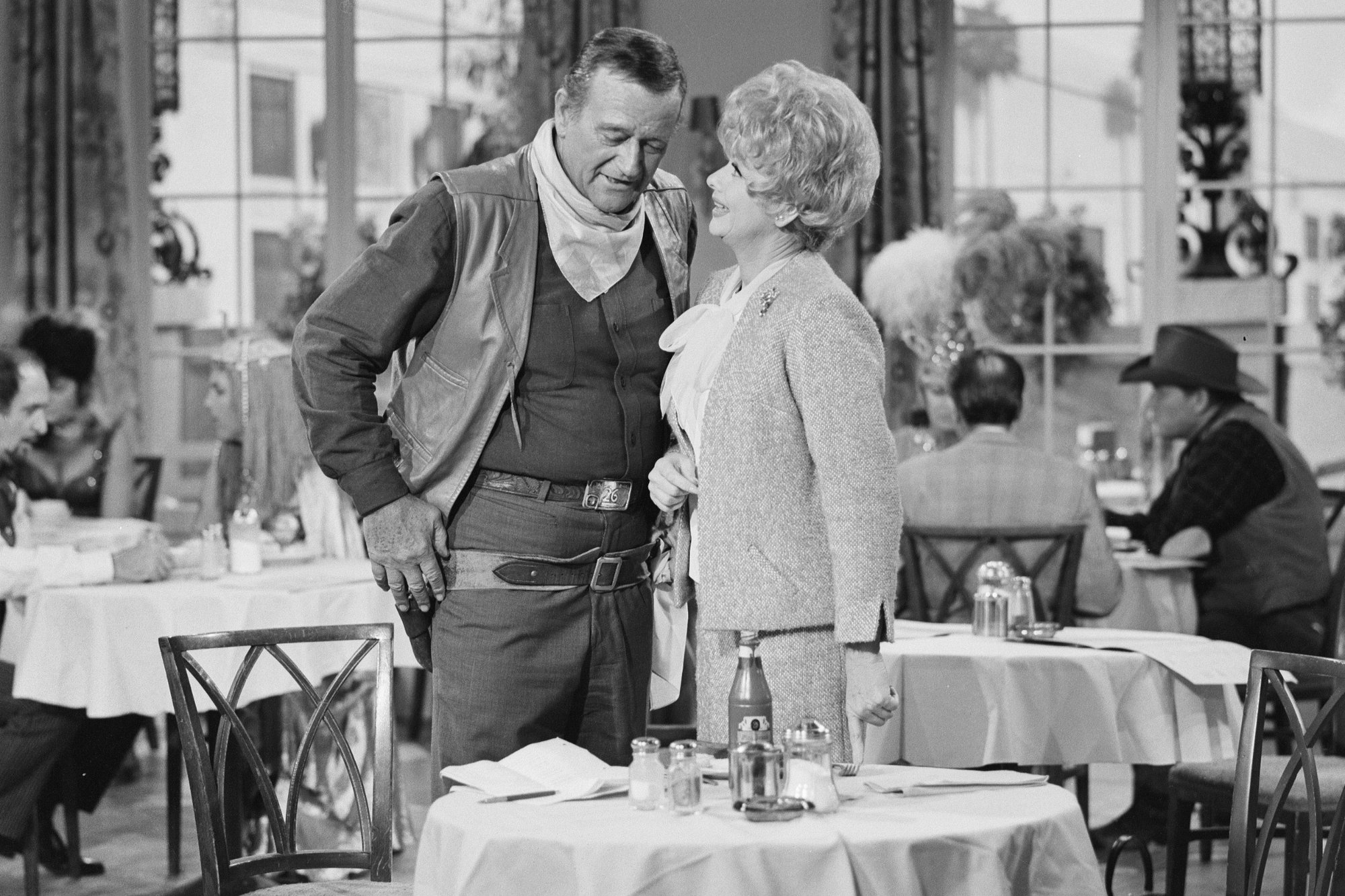 'The Lucy Show' John Wayne as himself and Lucille Ball as Lucy Carmichael. Black-and-white photo of Wayne wearing a cowboy outfit and Ball wearing a jacket and skirt. They're standing behind a table at a restaurant.