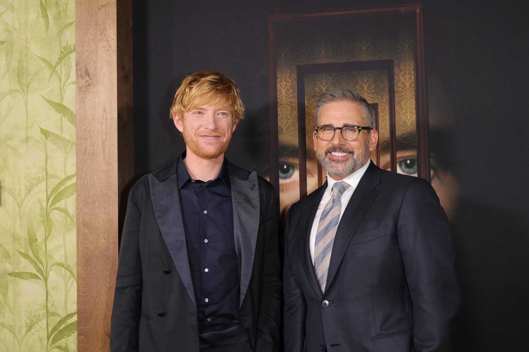 Domhnall Gleeson and Steve Carell on the red carpet for 'The Patient.' They're standing in front of key art for the series, wearing suits, and smiling.