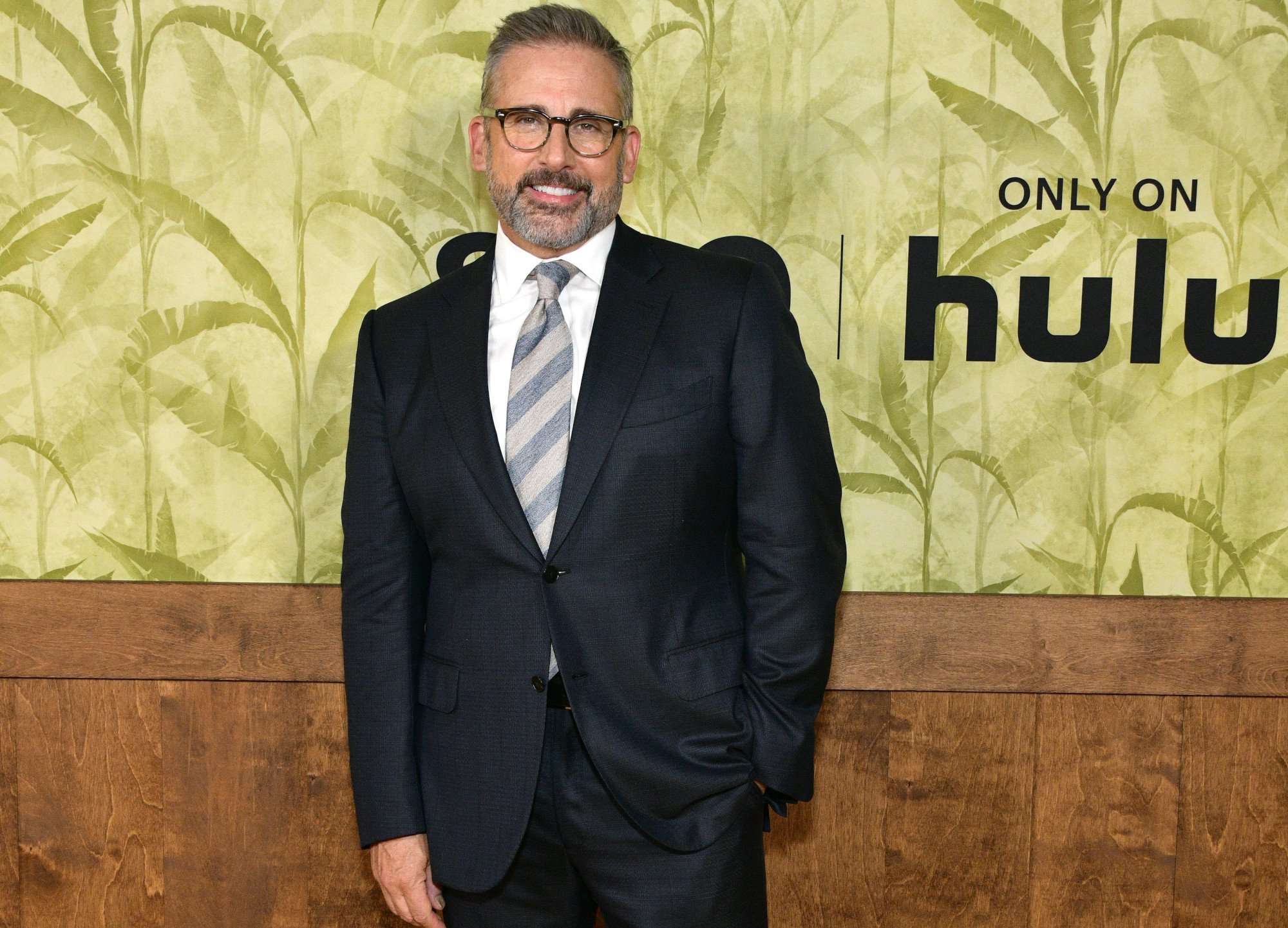 Steve Carell at the premiere for 'The Patient.' He's wearing a black suit and grey striped tie, and he's standing in front of a gold and brown wall that says 'Hulu' on it.
