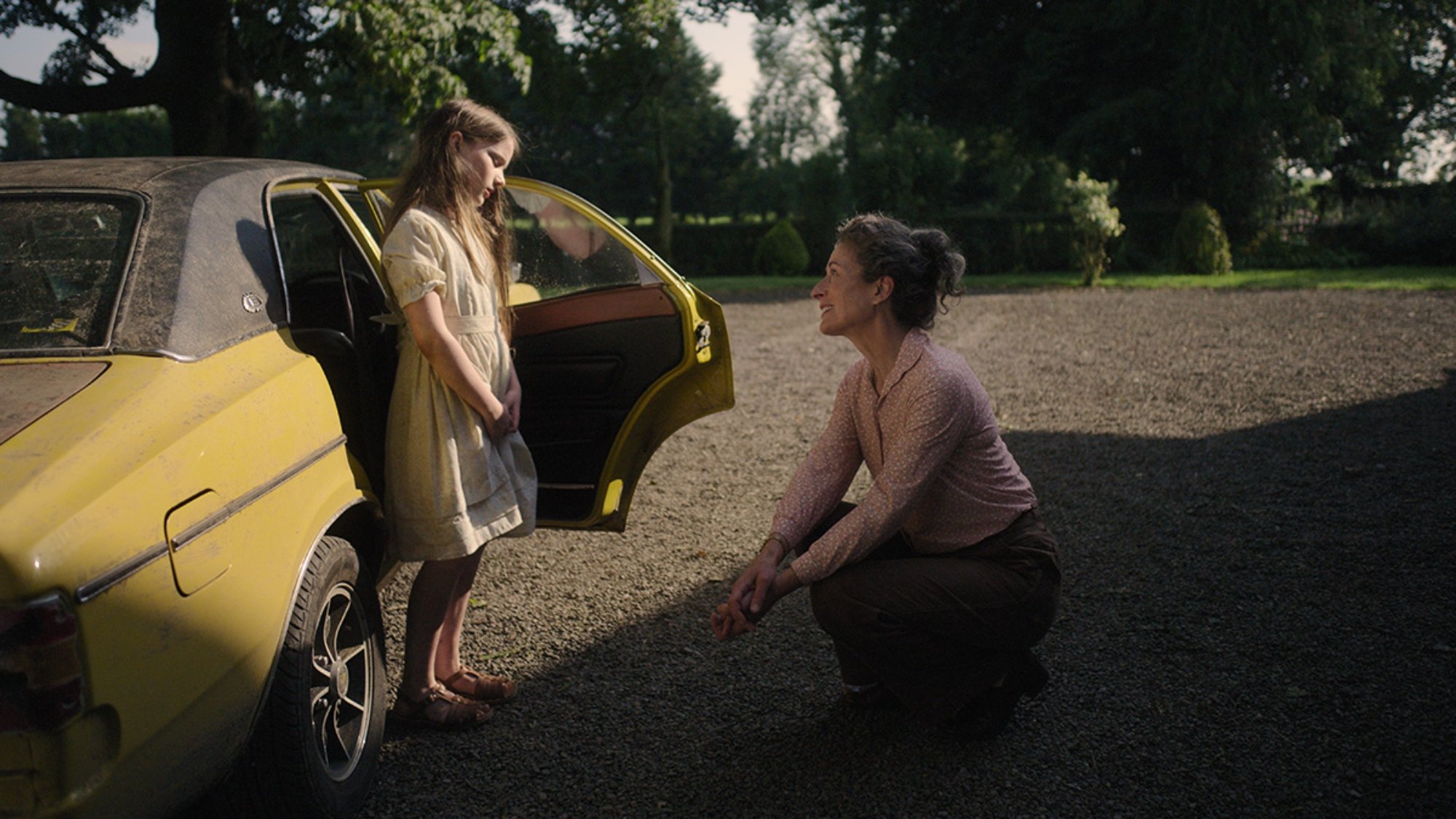 'The Quiet Girl' Catherine Clinch as Cáit and Carrie Crowley as Eibhlín Cinnsealach. Clinch is standing outside of a yellow car with the door open. Crowley is on her knees looking up at Clinch.