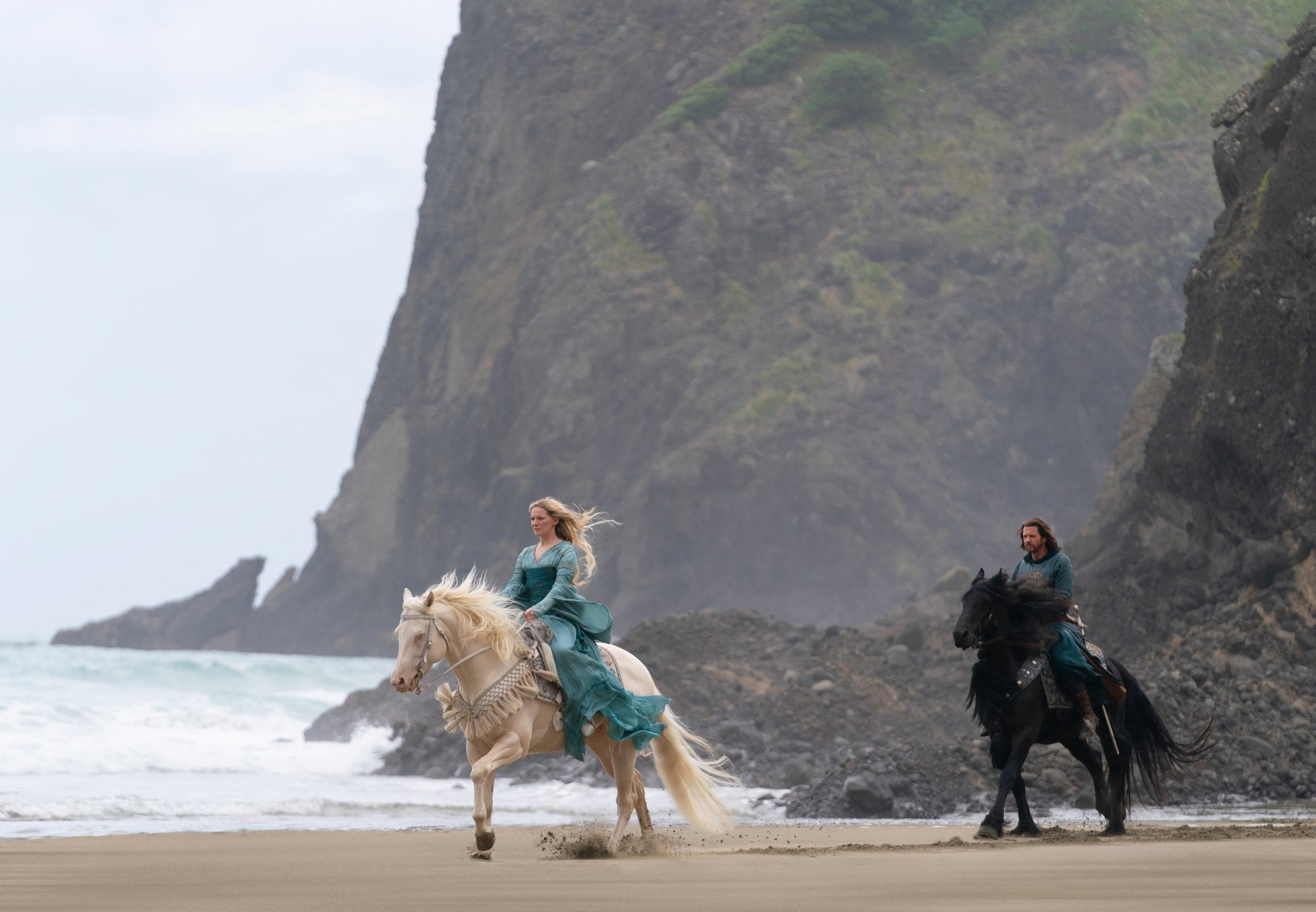 Morfydd Clark and Lloyd Owen as Elendil in 'The Lord of the Rings: The RIngs of Power,' which releases episode 3 on Sept. 9. They're riding horses in front of a cliff and the shore.