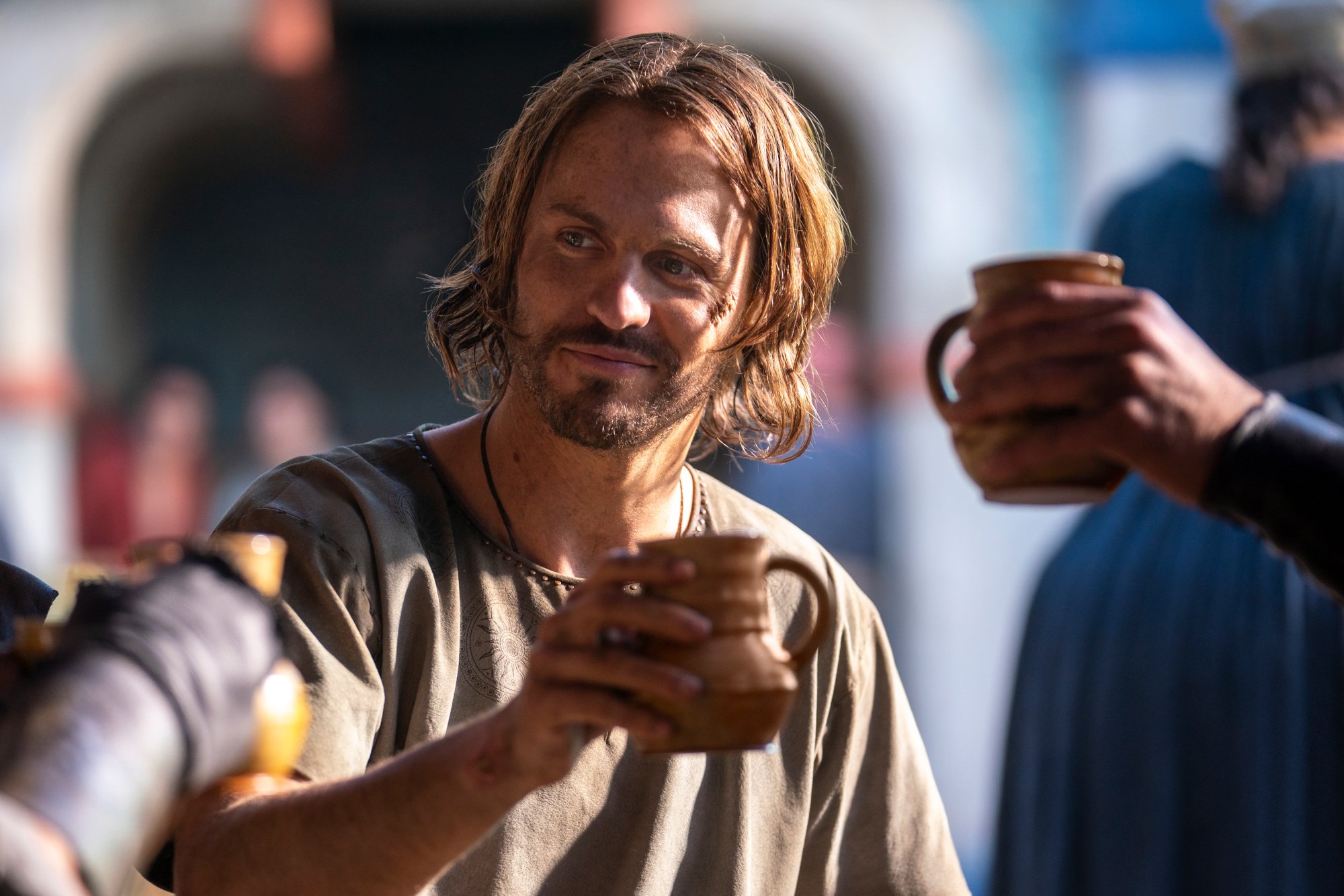 Charlie Vickers as Halbrand in 'The Lord of the Rings: The Rings of Power.' He's holding a mug and smiling.