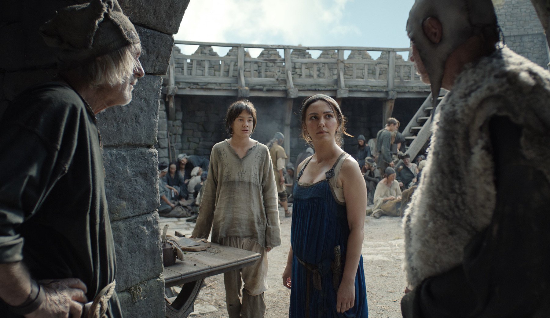 Tyroe Muhafidin and Nazanin Boniadi as Theo and Bronwyn in 'The Rings of Power,' where they're both running from Orcs in the Southlands. In the image, they're talking to two men and Bronwyn looks unhappy with them.