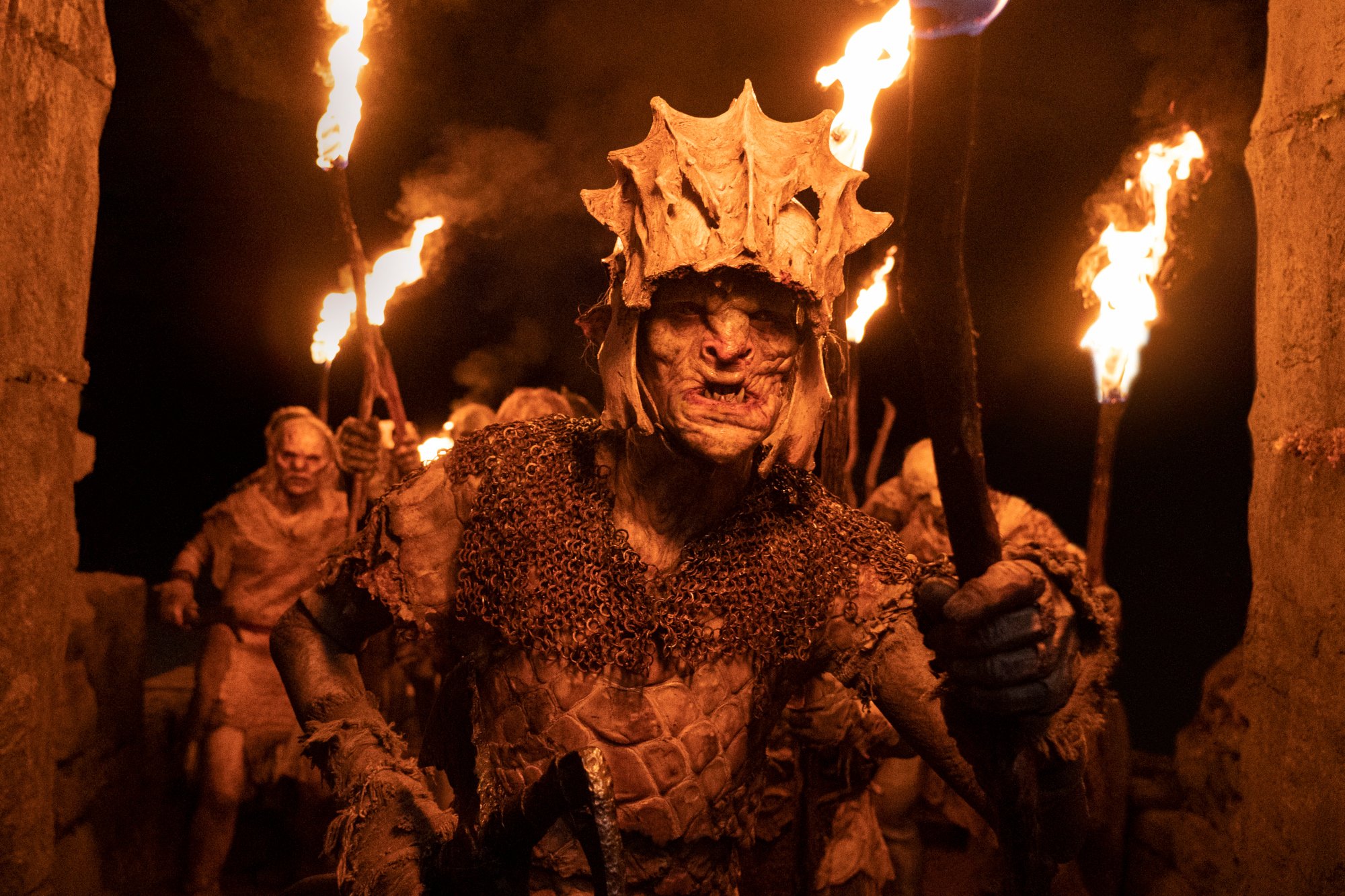 The Orcs following the orders of Adar in 'The Lord of the Rings: The Rings of Power.' They're holding torches and the leader is wearing a helmet.