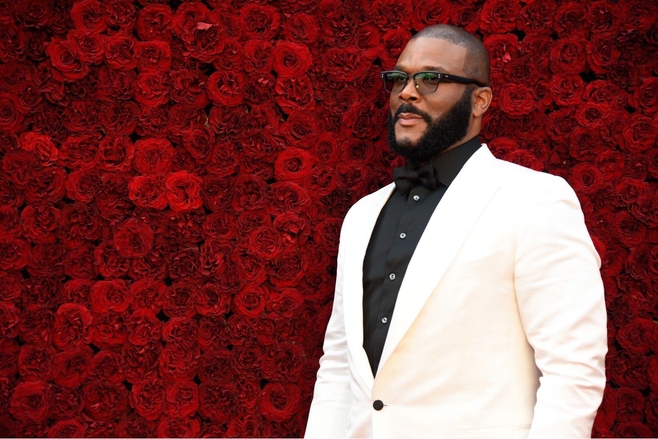 Tyler Perry wears a white jacket and black shirt and stands in front of a flower wall.
