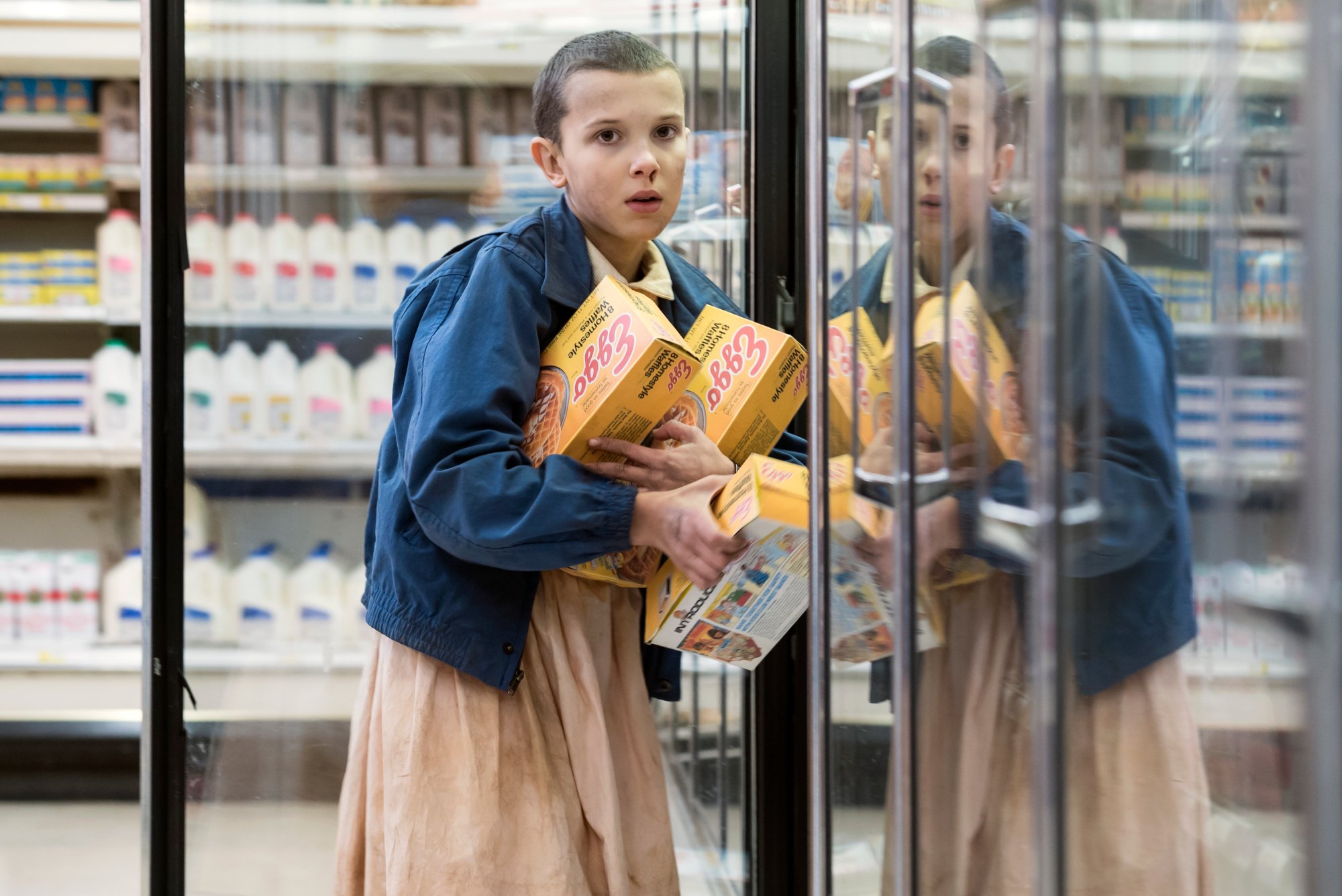 Millie Bobby Brown as Eleven in 'Stranger Things' Season 1 for our article about where to watch the show. She's standing next to a store refrigerator and holding several boxes of Eggo waffles.