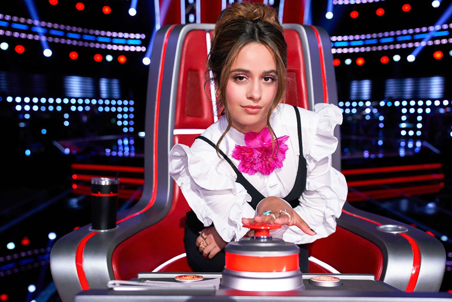 Camila Cabello poses in her chair with her hand on her button on The Voice Season 22