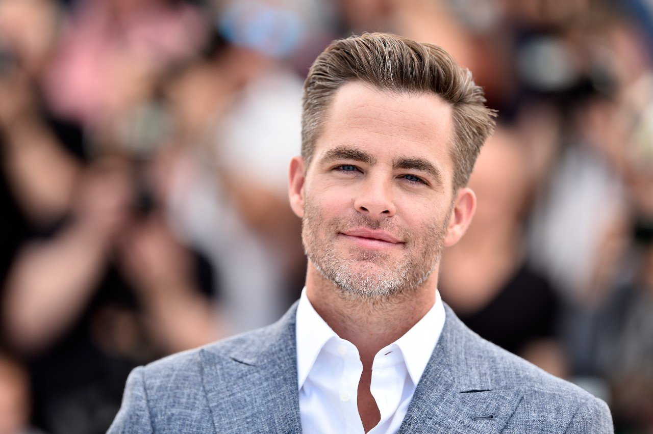 Chris Pine smiles while wearing a white shirt and grey suit jacket.