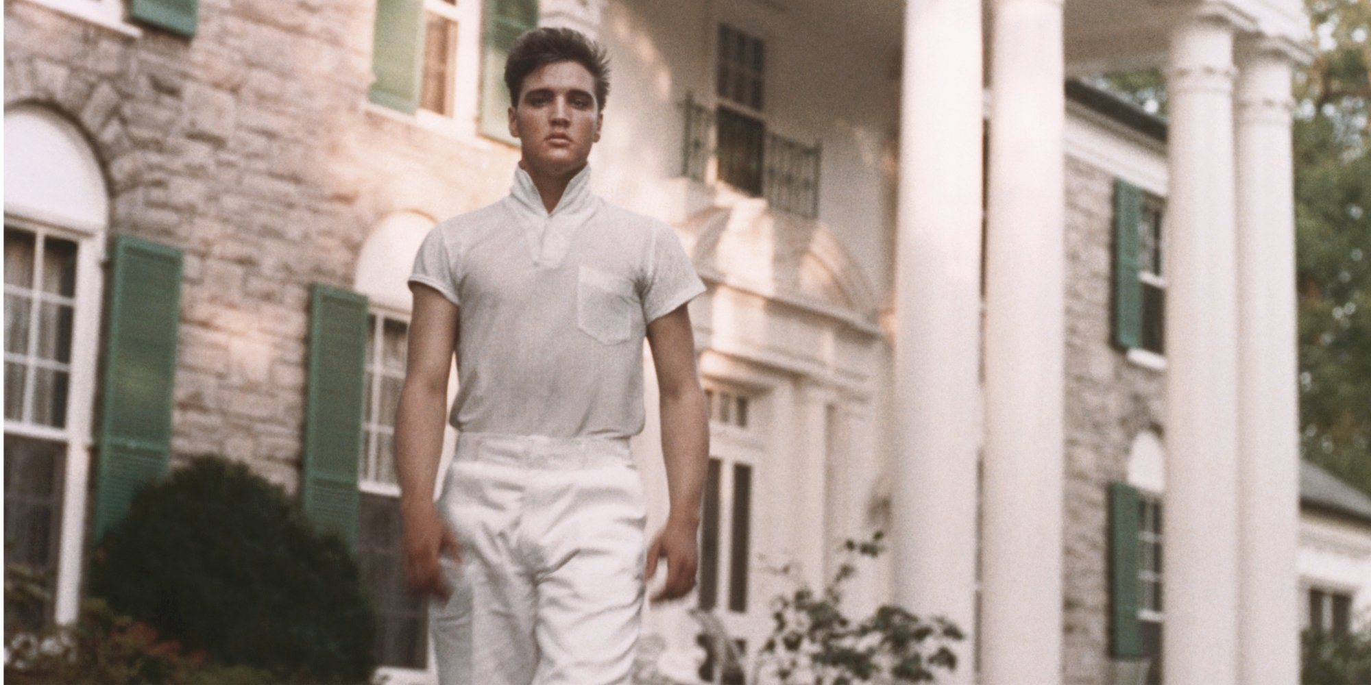 Elvis Presley poses in front of Graceland in a color photograph.