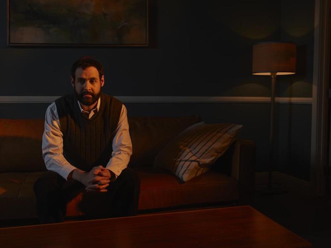 Ezra (Andrew Leeds) sits on a couch in a commercial for The Patient