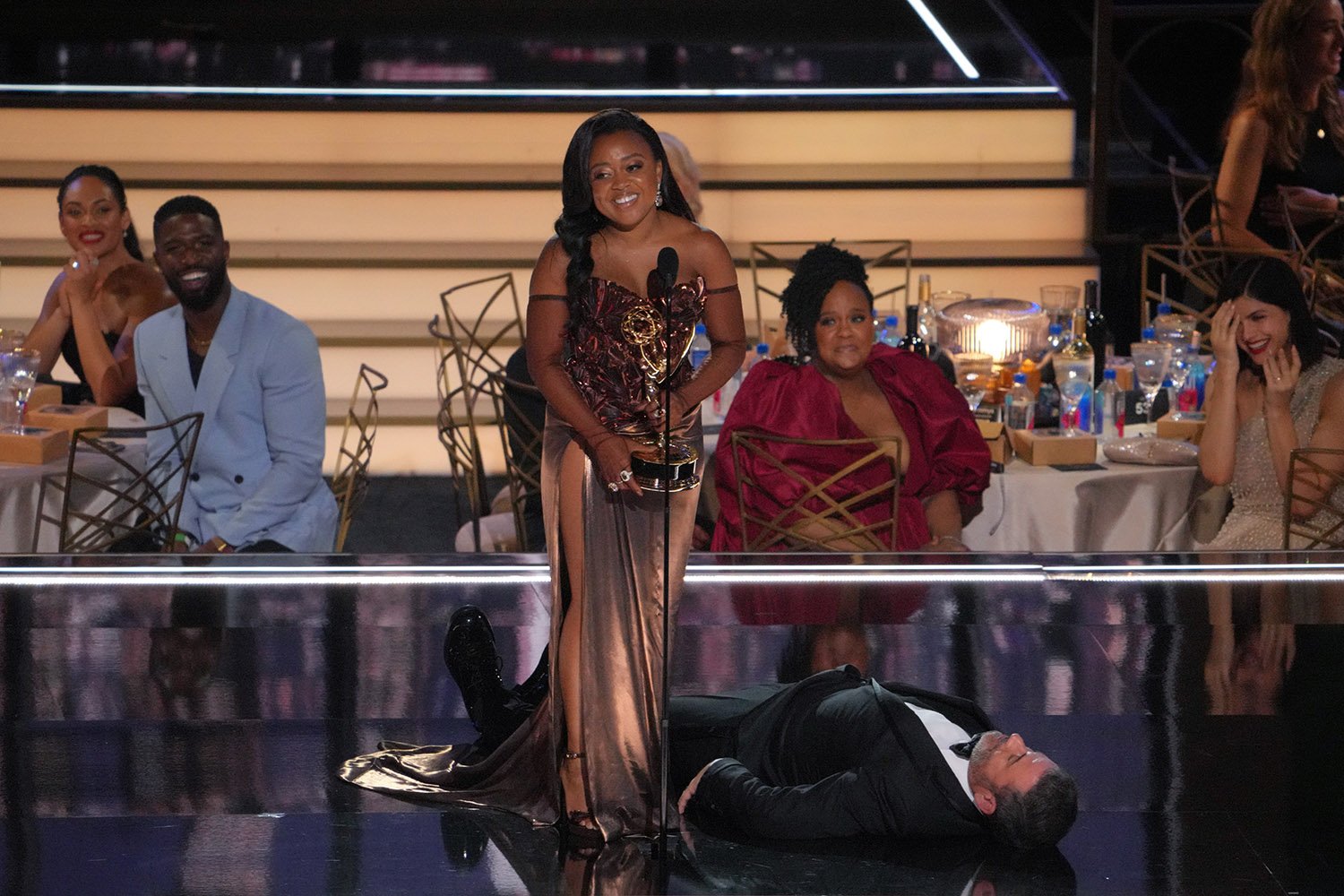 Quinta Brunson accepts her award at the Emmys 2022 as Jimmy Kimmel pretends to be passed out on stage beside her.