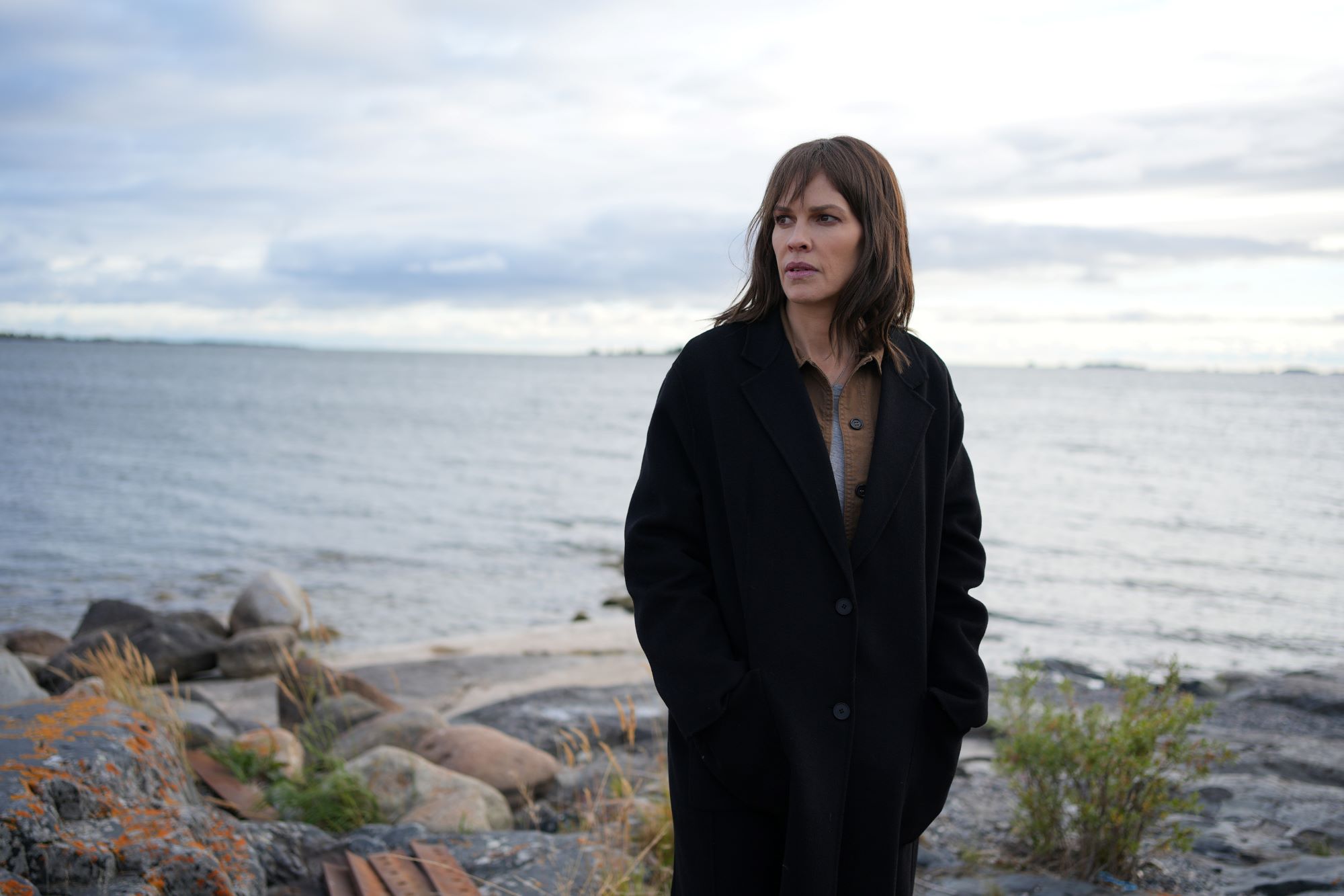 Hilary Swank, in character as Eileen Fitzgerald in the show 'Alaska Daily' on ABC, wears a black coat over a brown button-up shirt and stands in front of a body of water.