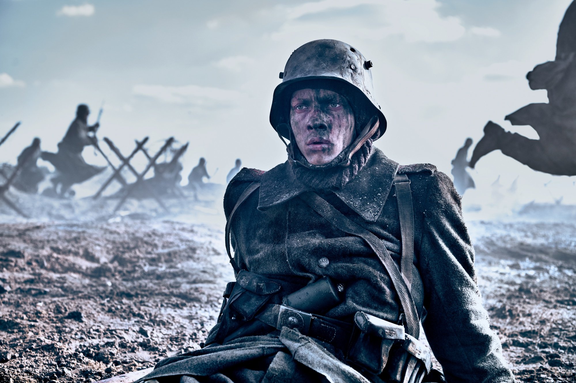 'All Quiet on the Western Front' Felix Kammerer as Paul Bäumer wearing his war uniform, sitting on the battlefield looking in terror with his face covered in mud. Other soldiers running in the background.