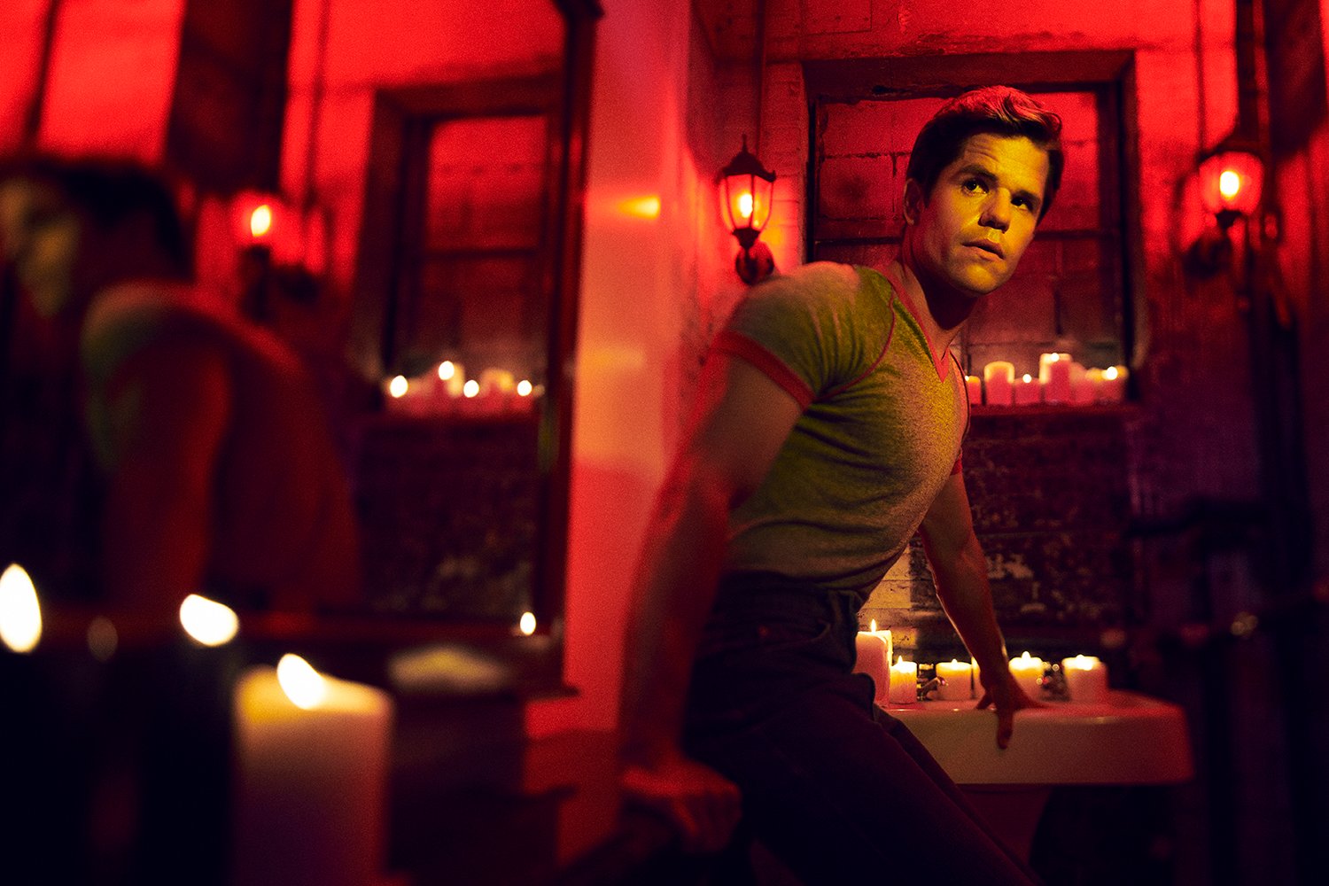 Charlie Carver as Adam, sitting on the floor of a red candlelit room in American Horror Story: NYC.