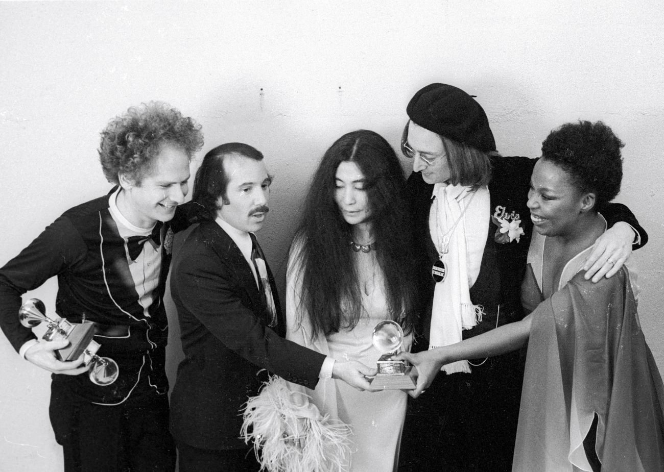 A black and white photo of Art Garfunkel, Paul Simon, Yoko Ono, John Lennon, and Roberta Flack hold Grammy awards.
