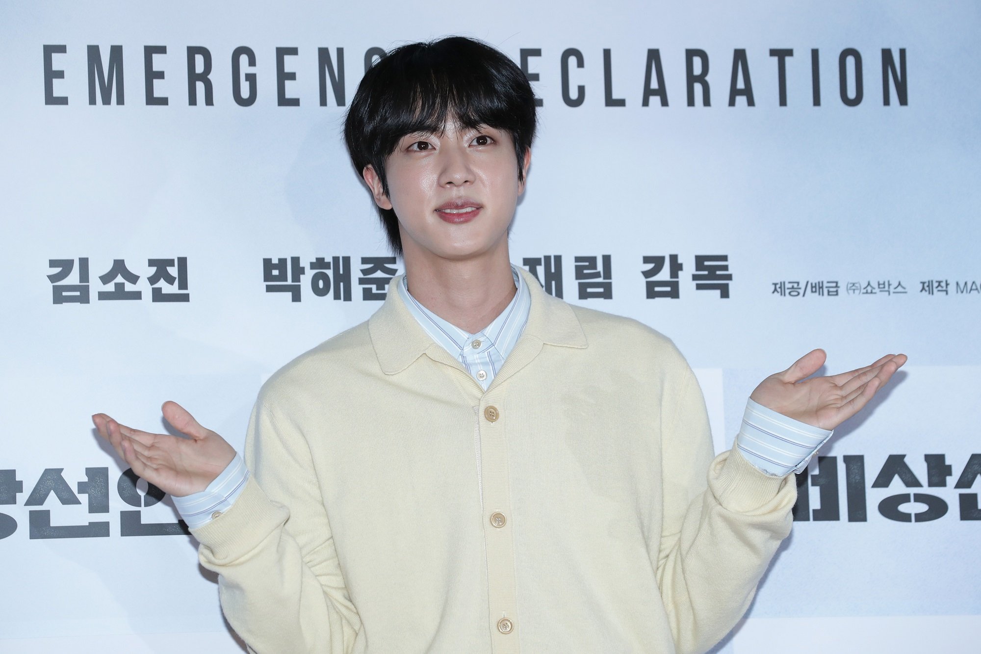 Jin of BTS shrugs while wearing a light yellow sweater