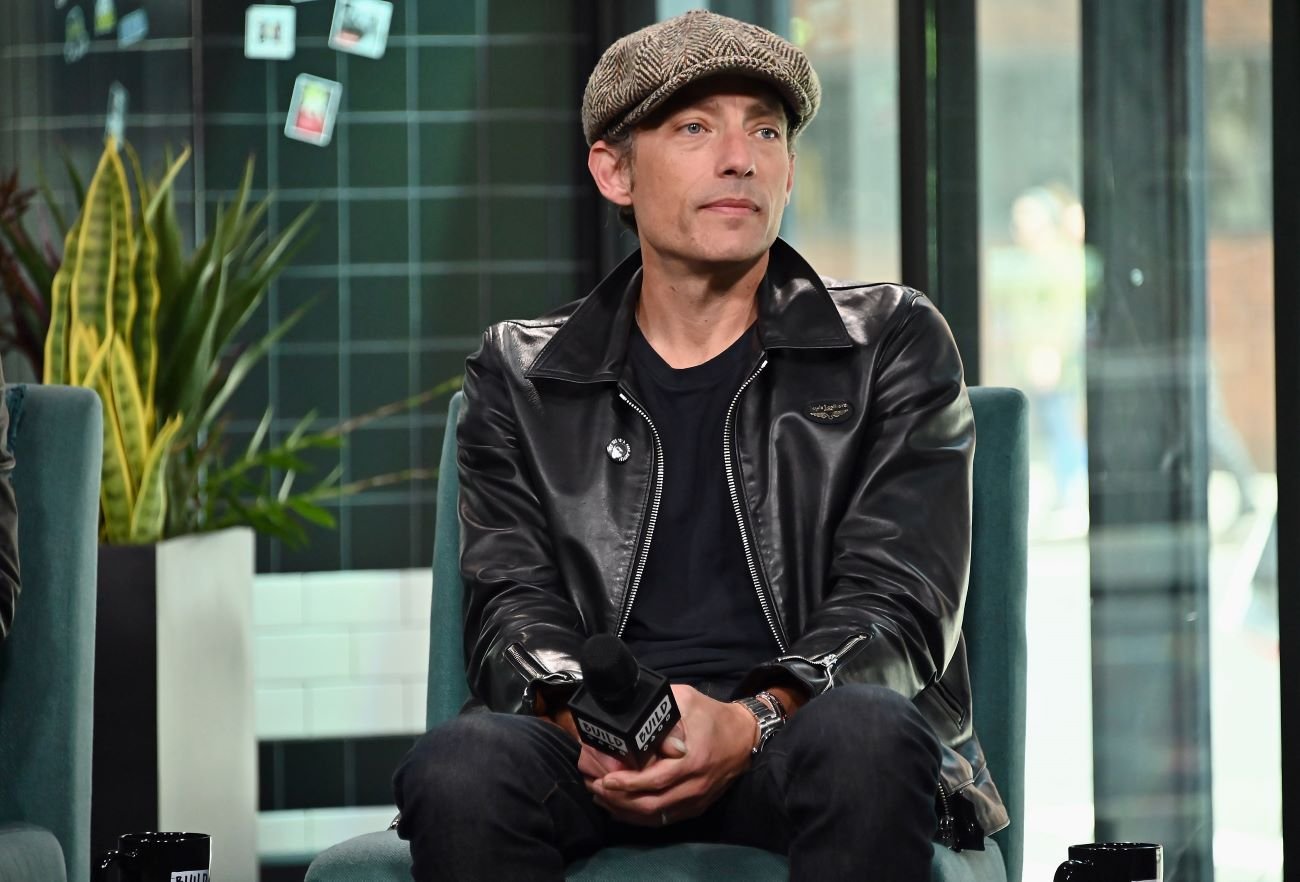Jakob Dylan wears a hat and sits in a chair.