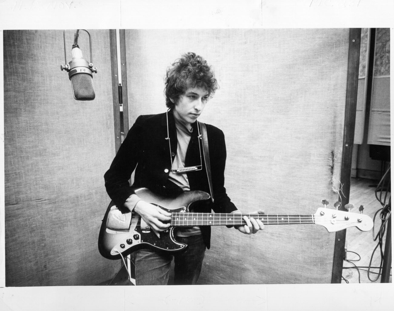 A black and white picture of Bob Dylan playing a guitar with a harmonica around his neck.