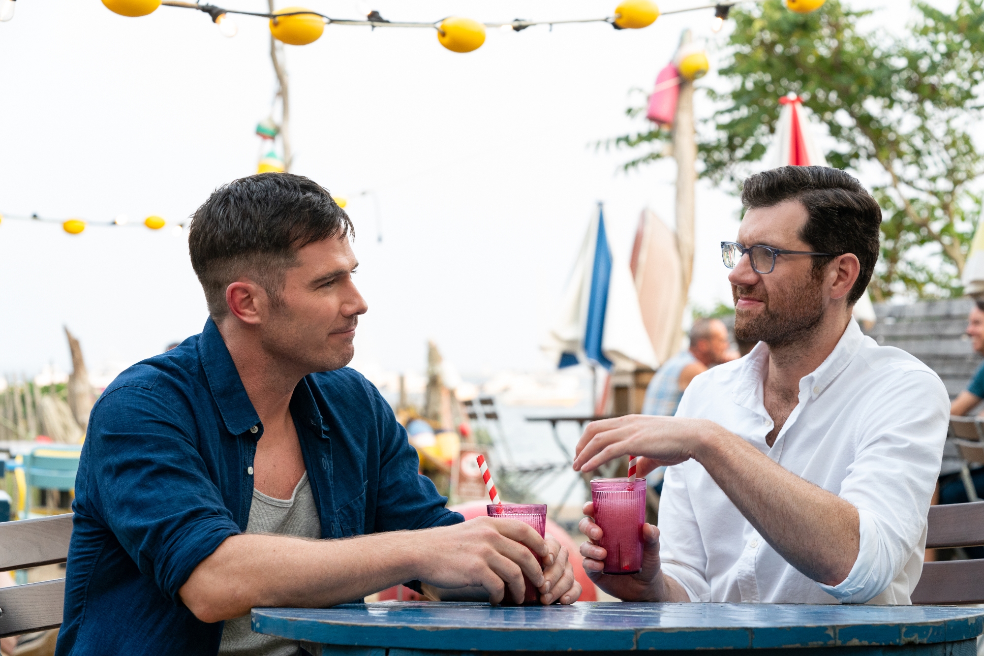 'Bros' Luke Macfarlane as Aaron and Billy Eichner as Bobby in box office disaster. They're sitting at a table holding their drinks in bright-colored glasses with white and red straws.