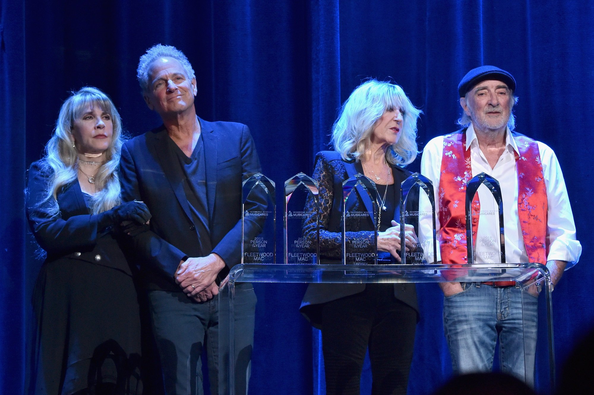 Stevie Nicks stands arm-in-arm with Lindsey Buckingham beside Christine McVie and John McVie on stage in front of a blue curtain