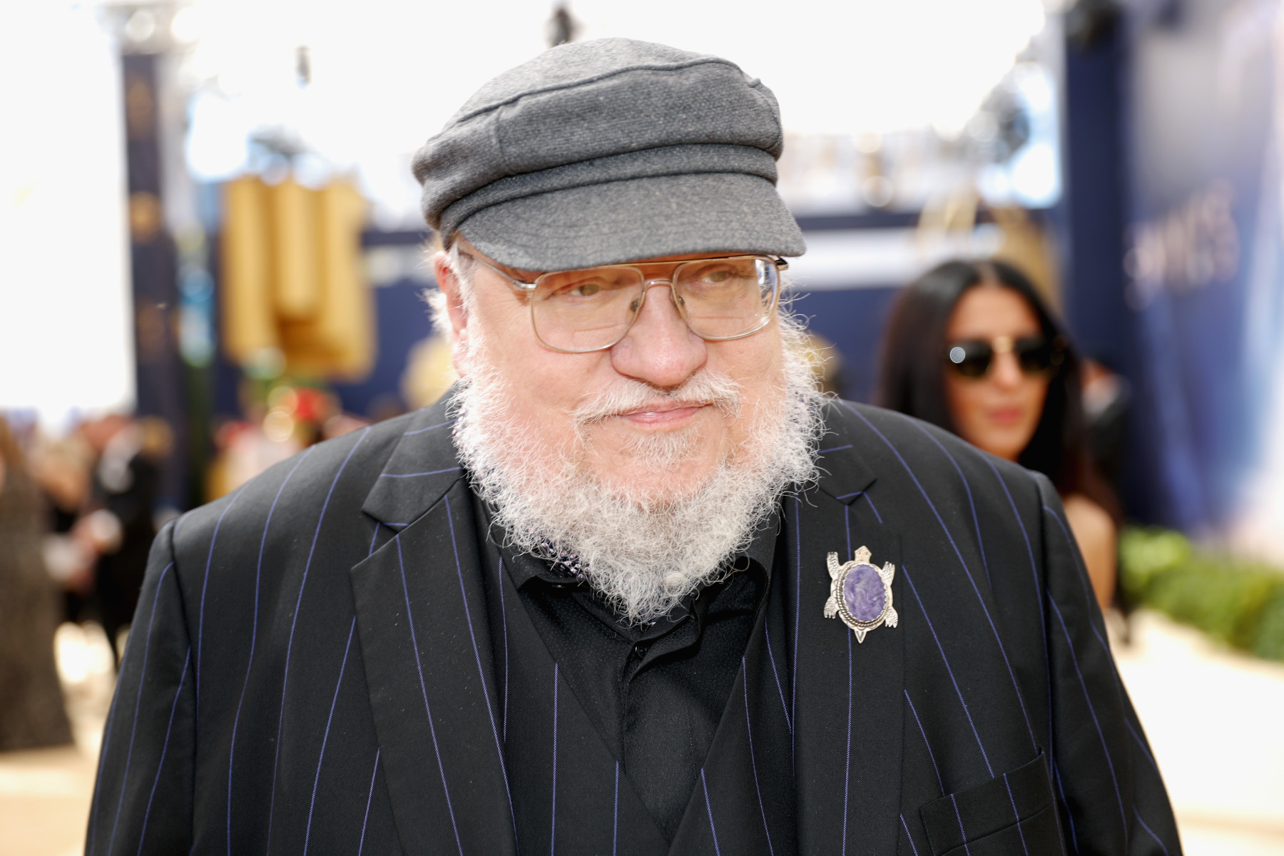George R.R. Martin, who addressed the rivalry between 'House of the Dragon' and 'The Rings of Power.' He's wearing a grey hat, black suit, and pin on his left side.