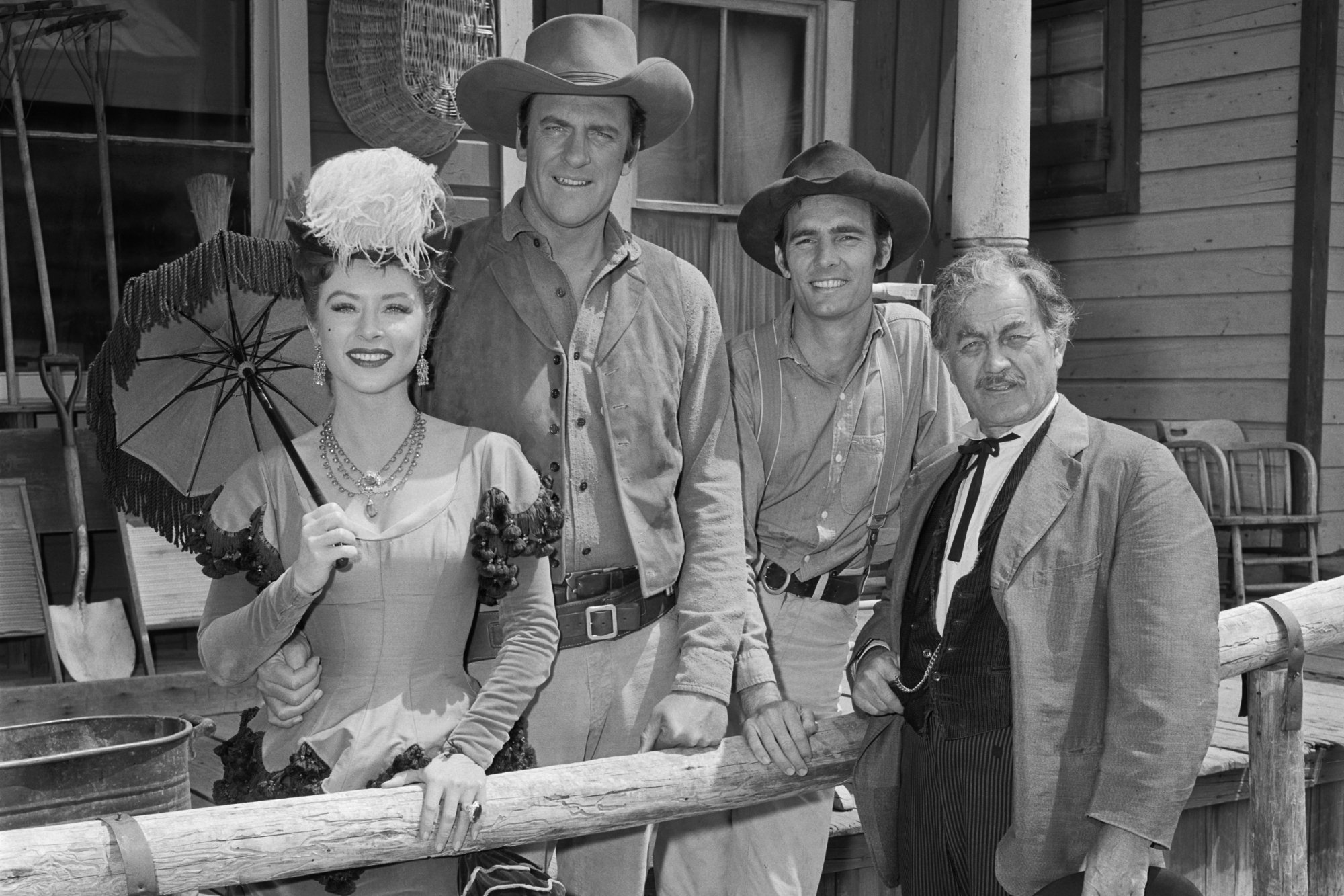 'Gunsmoke' Amanda Blake as Kitty Russell, James Arness as Marshal Matt Dillon, Dennis Weaver as Chester Goode and Milburn Stone as Dr. Galen 'Doc' Adams in best episodes. They're standing behind wooden fence, smiling.