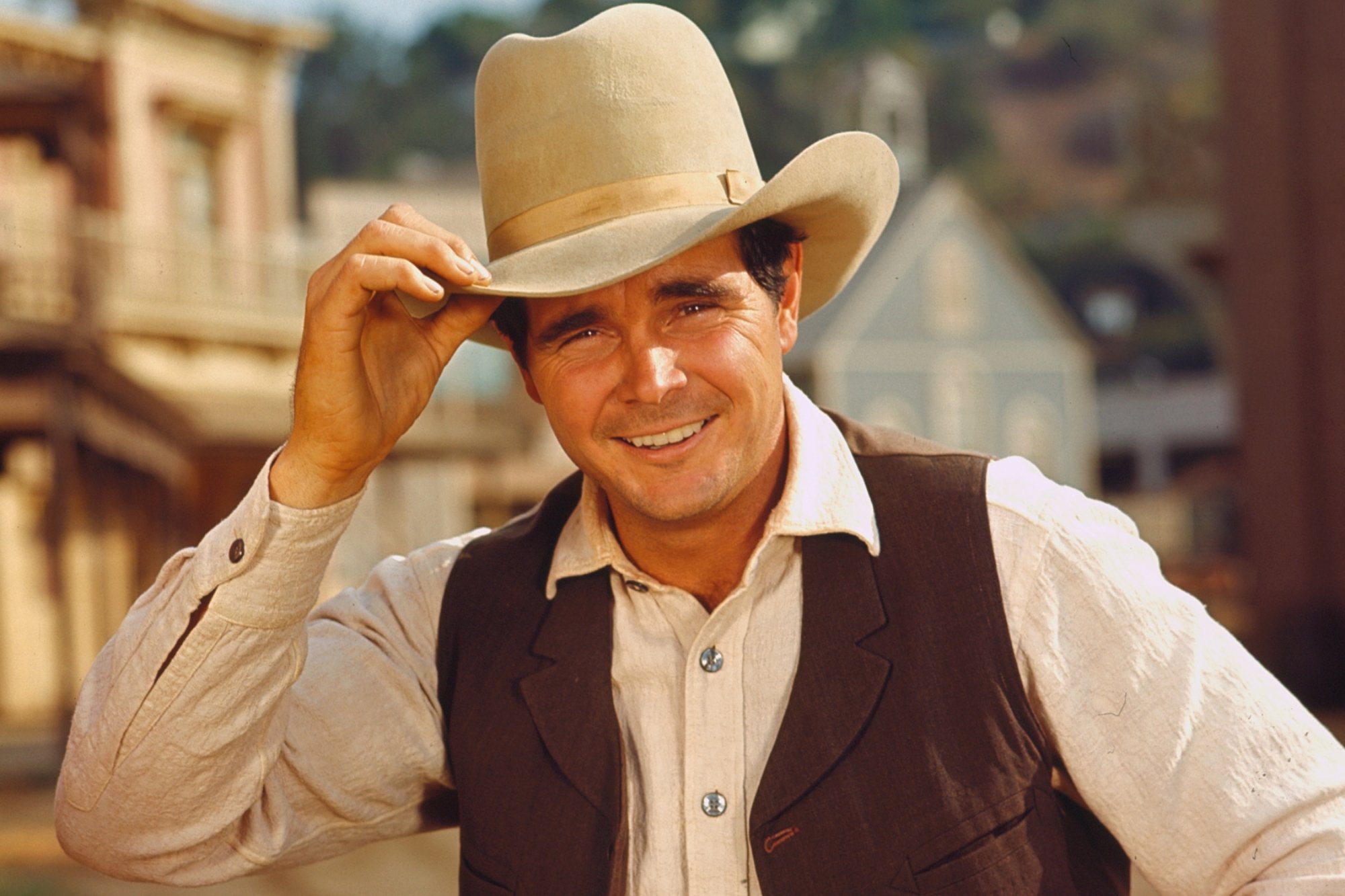 'Gunsmoke' Buck Taylor as Newly O'Brien wearing a vest, collared shirt, and cowboy hat. He has his hand on the rim of the hat and is smiling.
