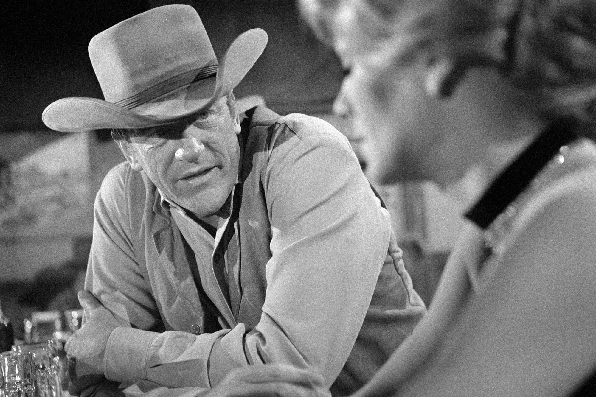 'Gunsmoke' James Arness as Matt Dillon and Betty Hutton as Molly McConnell, who had an episode perfect for Halloween. He's leaning on the counter, looking at her, wearing his iconic Western outfit with cowboy hat, vest, and collared shirt.