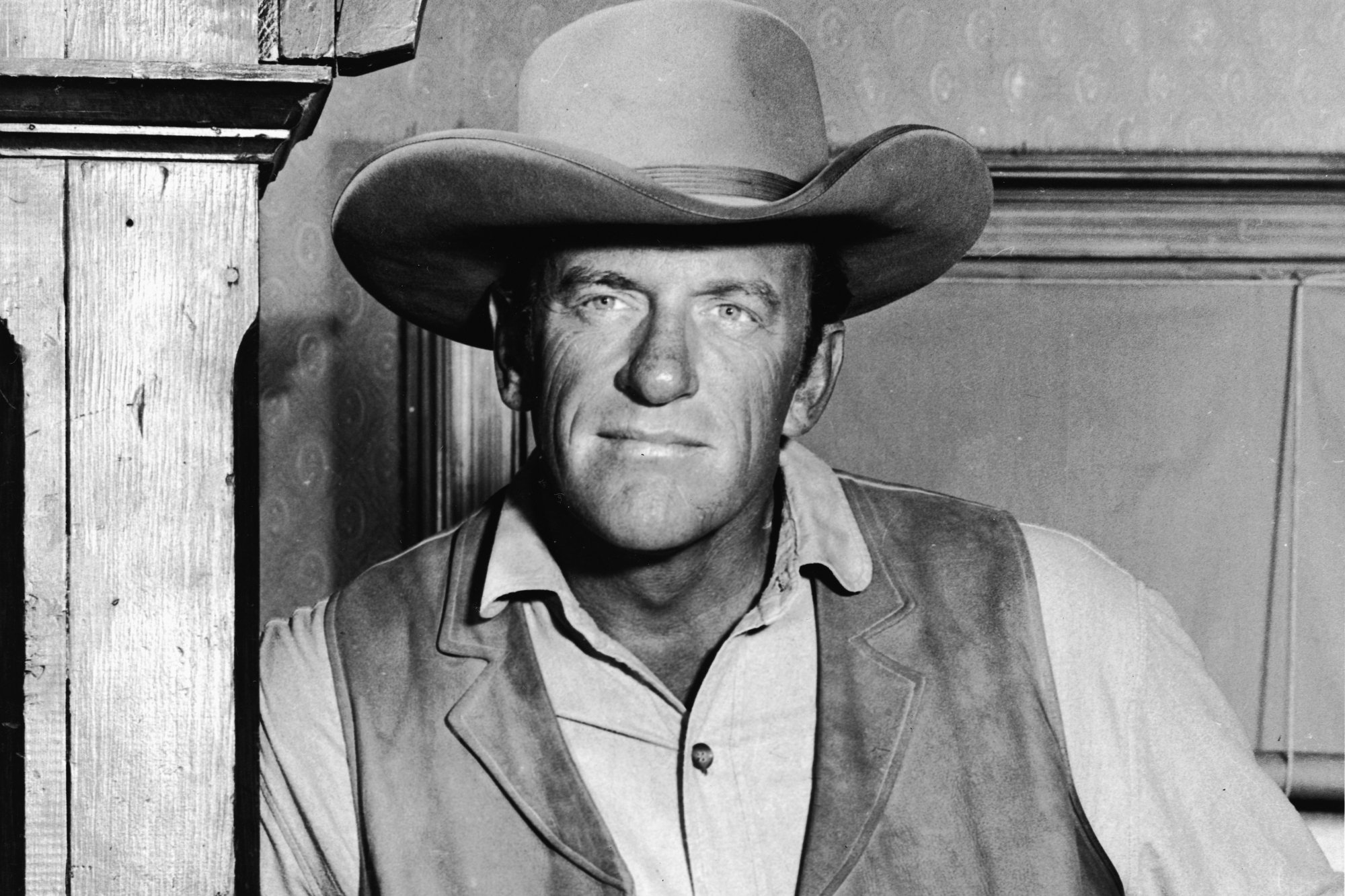 'Gunsmoke' James Arness as Matt Dillon wearing a vest, collared shirt, and a cowboy hat. He's leaning against a pillar.