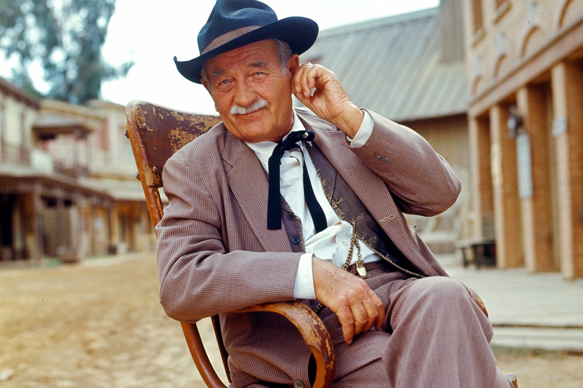 'Gunsmoke' Milburn Stone as Doc Adams sitting in a wooden chair wearing a vest, pants, and Western hat. He's touching his earlobe.
