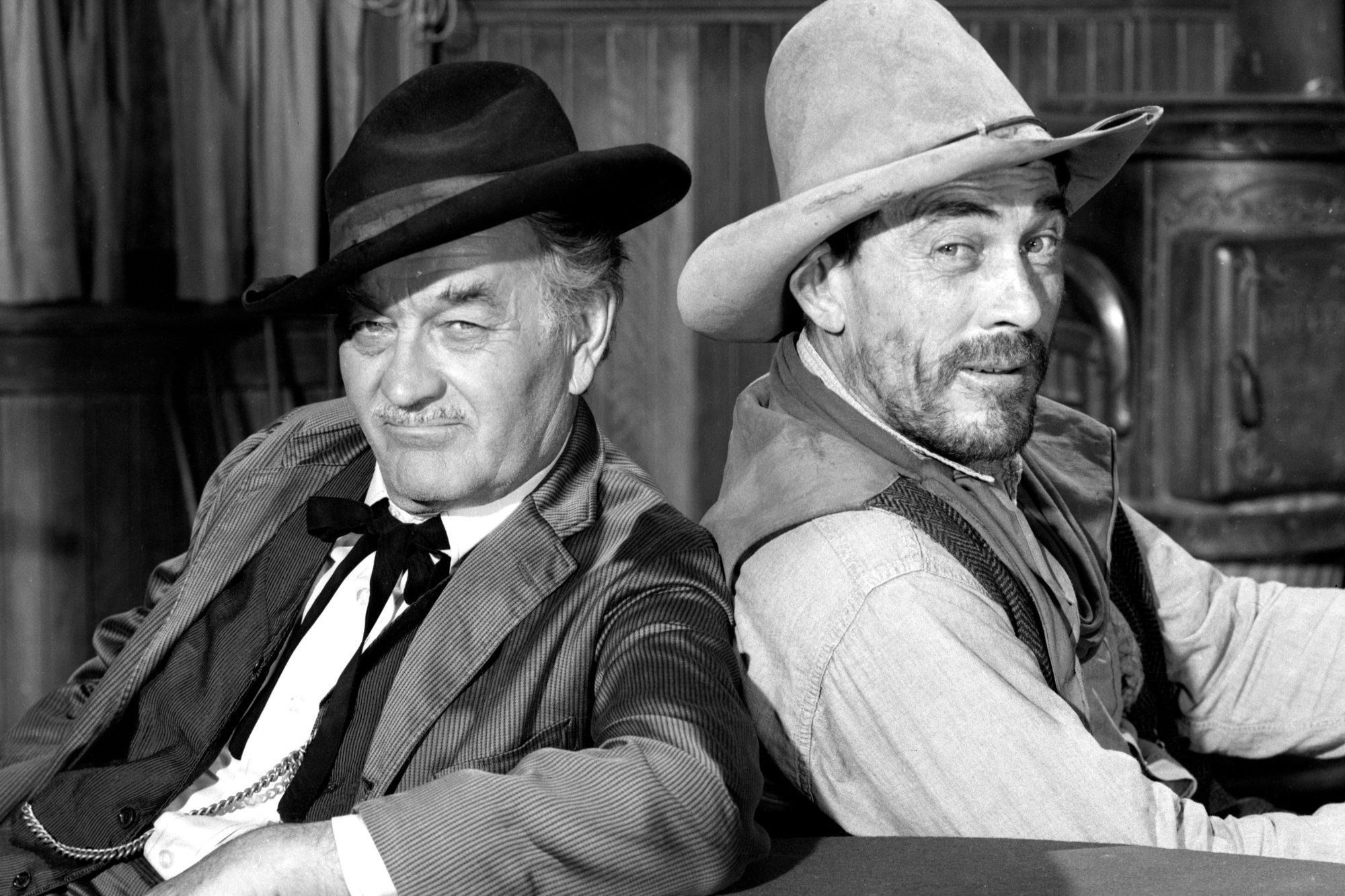 'Gunsmoke' Milburn Stone as Doc Adams and Ken Curtis as Festus Haggen leaning against one another with their shoulders toward one another. They're wearing Western costumes with slight smiles on their faces in a black-and-white photograph.