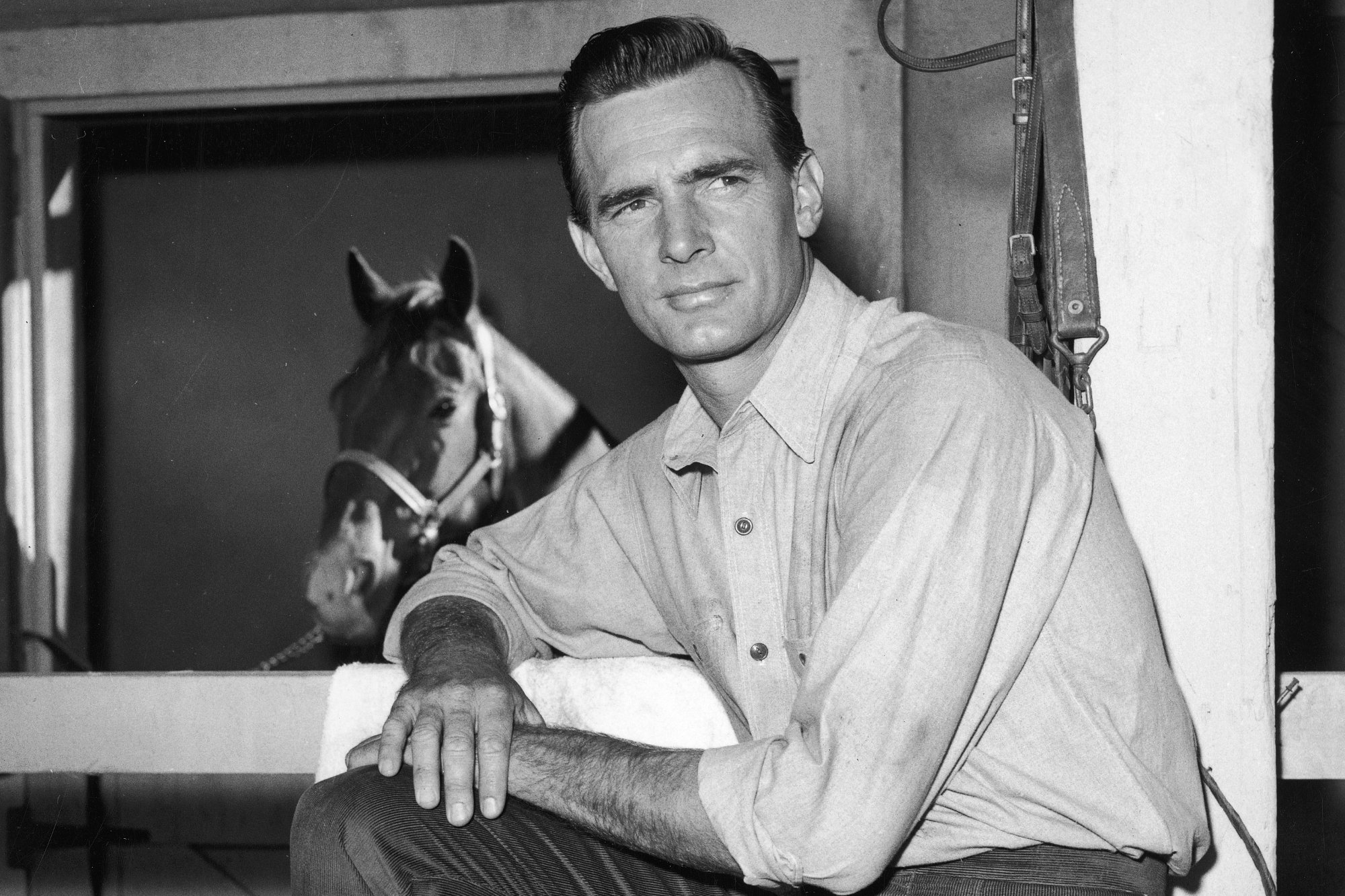 'Gunsmoke' actor Dennis Weaver wearing a collared shirt and resting his arms on a wooden gate with a horse in the background.
