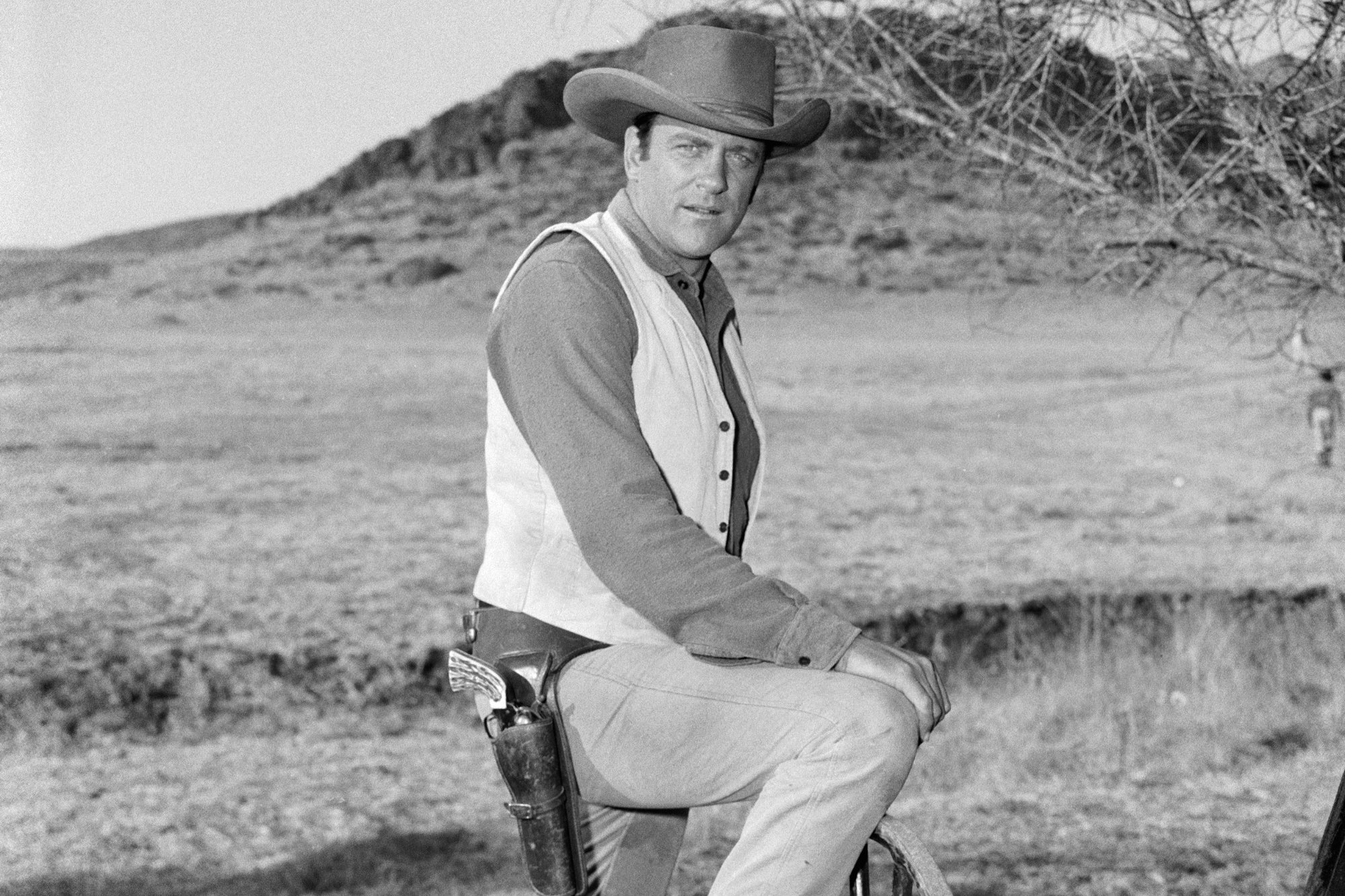 'Gunsmoke' actor James Arness as Marshal Matt Dillon in a black-and-white photograph in Western clothes with his foot resting on a carriage wheel.