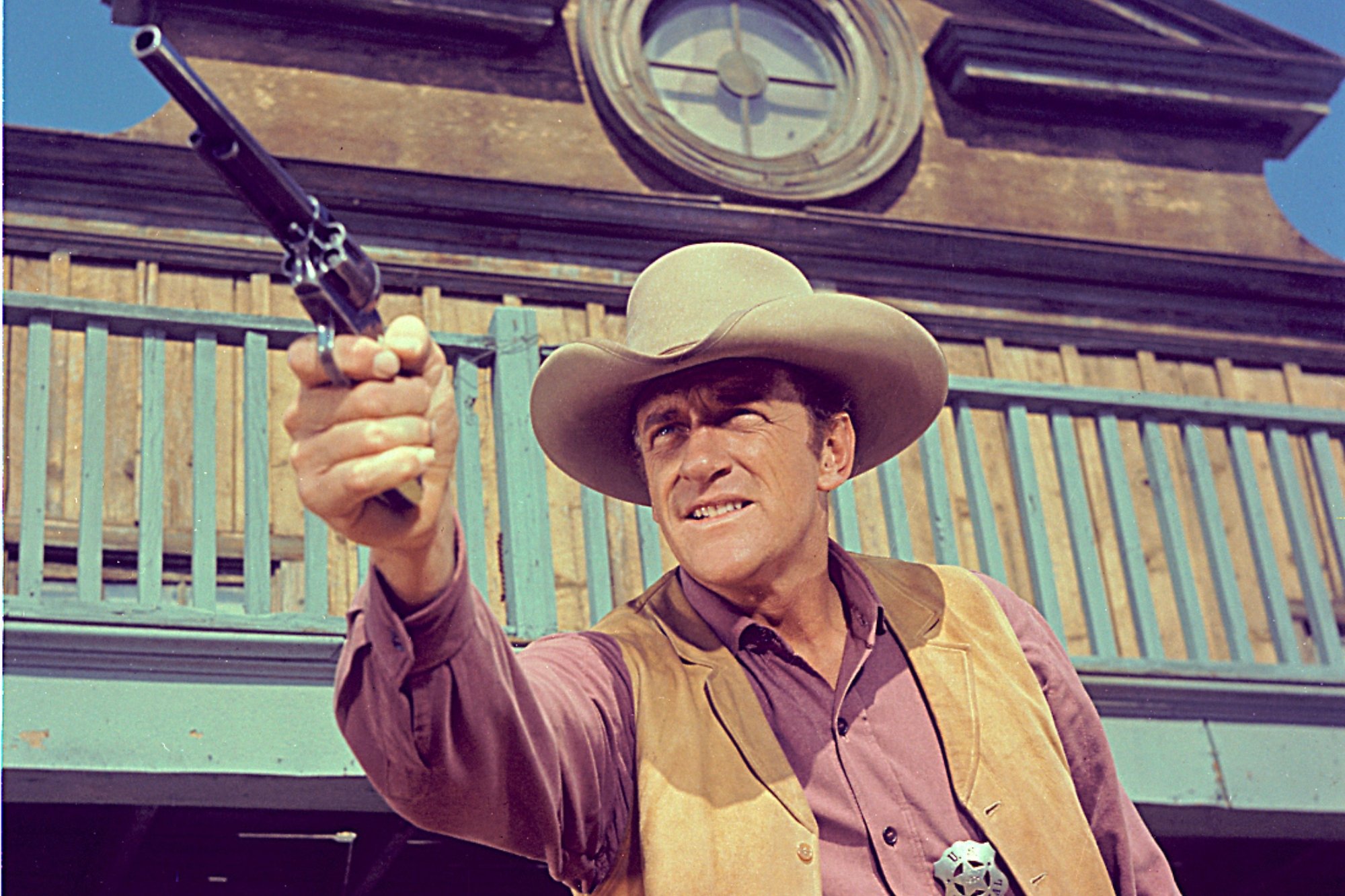 'Gunsmoke' actor James Arness as U. S. Marshal Matt Dillon pointing his gun, ready to shoot. He's wearing a tan vest and salmon-colored shirt underneath and a Western hat.