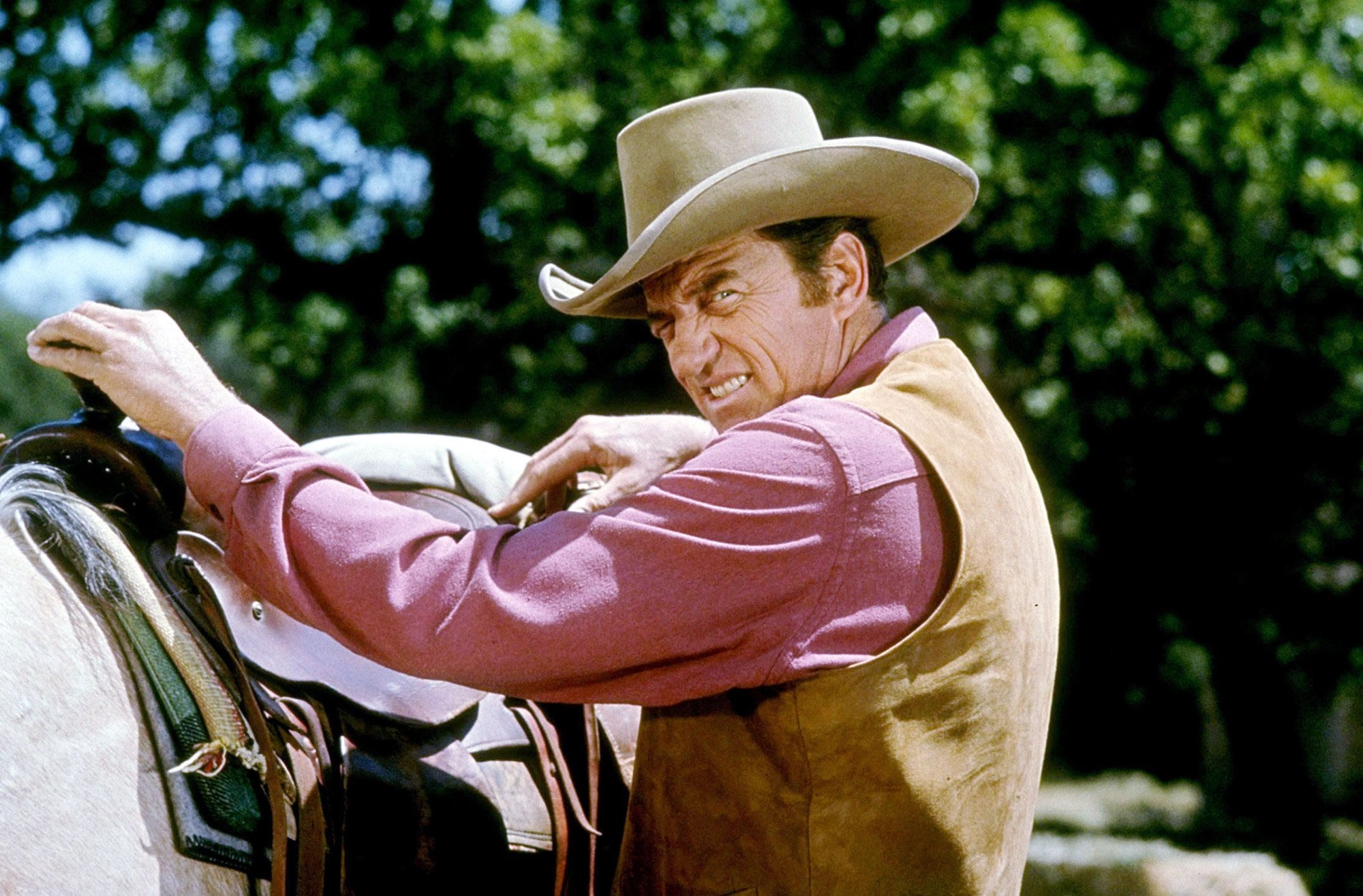 'Gunsmoke' actor James Arness as U.S. Marshal Matt Dillon with his hands on the horse saddle. His eyes are squinted, as he's looking over his shoulder wearing a tan vest and salmon shirt