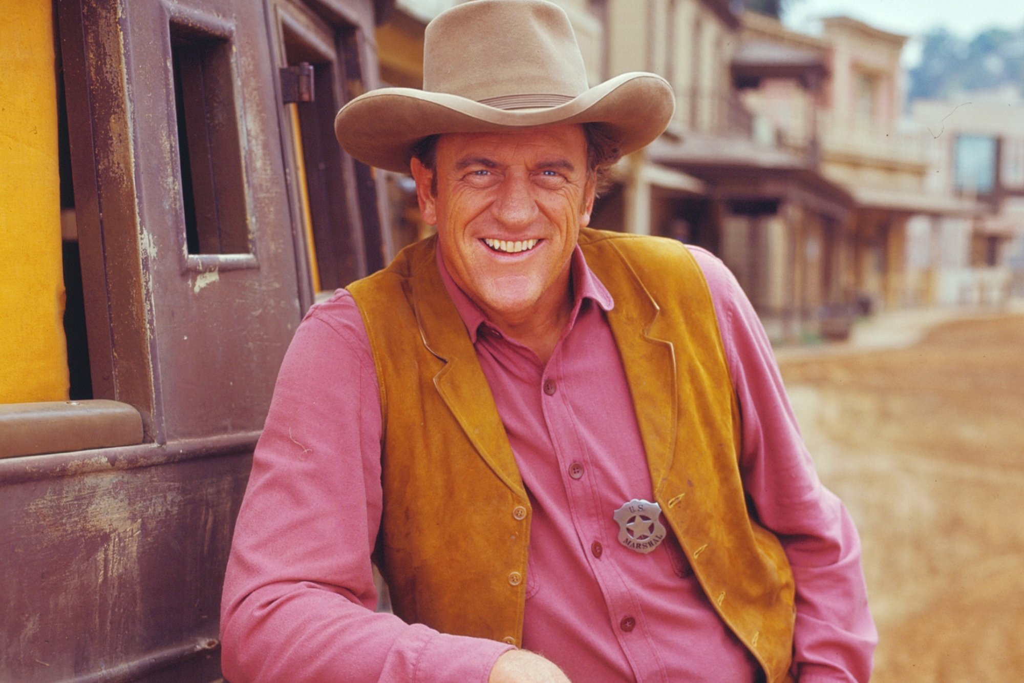 'Gunsmoke' actor James Arness as U. S. Marshal Matt Dillon leaning on carriage wearing a light tan vest and a pink-ish shirt. He has his badge on the shirt and is smiling.