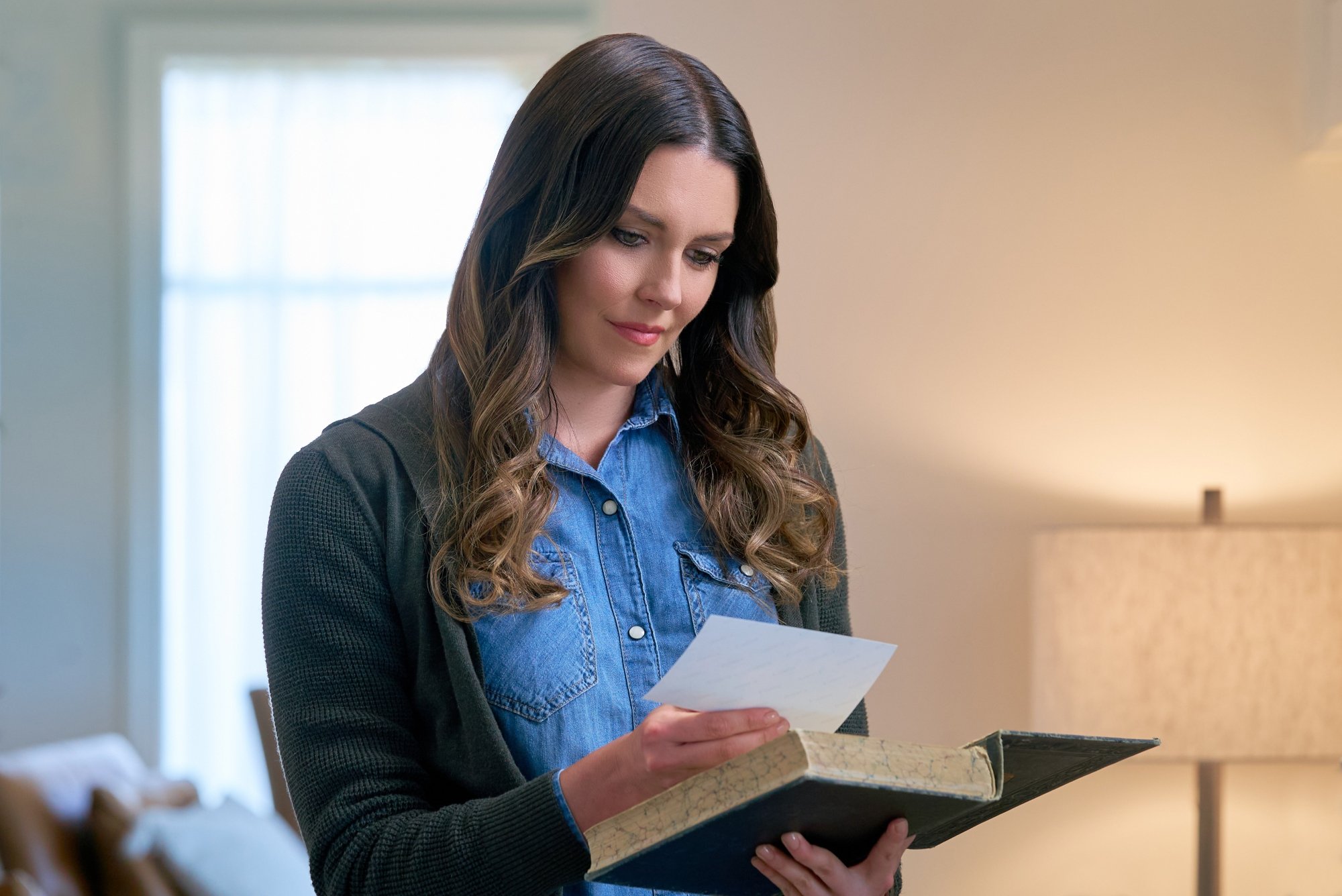 Hallmark's 'Pumpkin Everything' Taylor Cole as Amy wearing a denim-buttoned top. She's looking at a piece of paper she pulled from an open book.