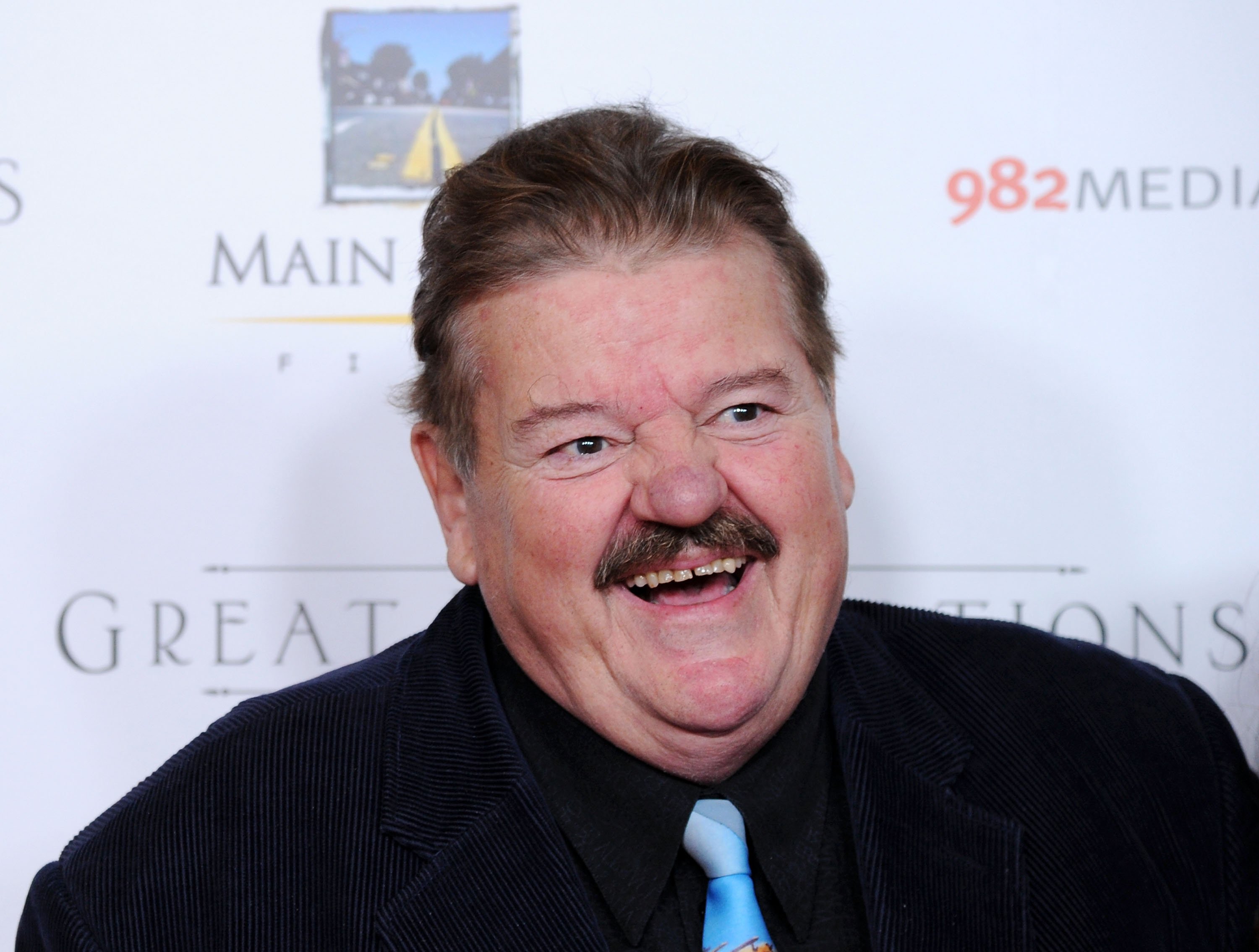 Robbie Coltrane, who starred in the 'Harry Potter' movies, wears a black suit over a black button-up shirt and light blue tie.